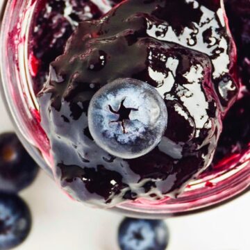 Close-up of a dessert jar topped with 3-Ingredient Blueberry Jam and a single blueberry. The jar is filled with dark purple sauce, and a few whole blueberries are scattered around it on a white surface.