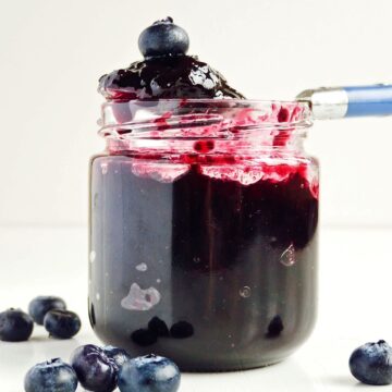 An open glass jar filled with thick 3-Ingredient Blueberry Jam, with a spoon dipped into the jar. The jam has some blueberries visible, and a few fresh blueberries are scattered around the base of the jar on a white surface.