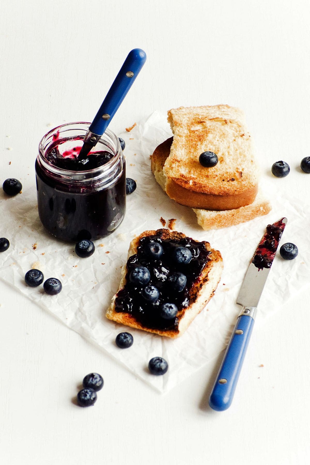 A jar of 3-Ingredient Blueberry Jam with a spoon inside sits on a piece of parchment paper. Beside it are two slices of toast. One slice is covered with the vibrant Refrigerator Jam and fresh blueberries, while the other slice is plain. A blue-handled knife and scattered blueberries are also visible.