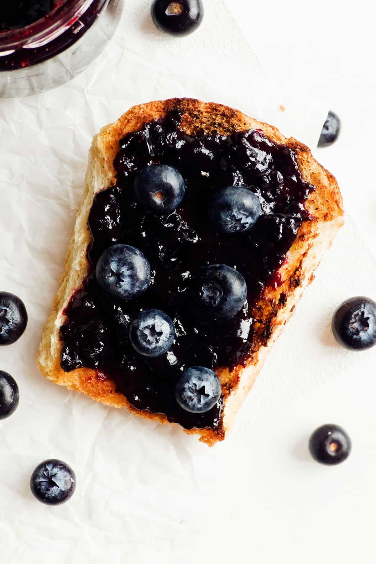 A slice of toasted bread topped with 3-ingredient refrigerator blueberry jam and fresh blueberries. The bread is placed on a piece of white parchment paper, and additional blueberries are scattered around the toast.