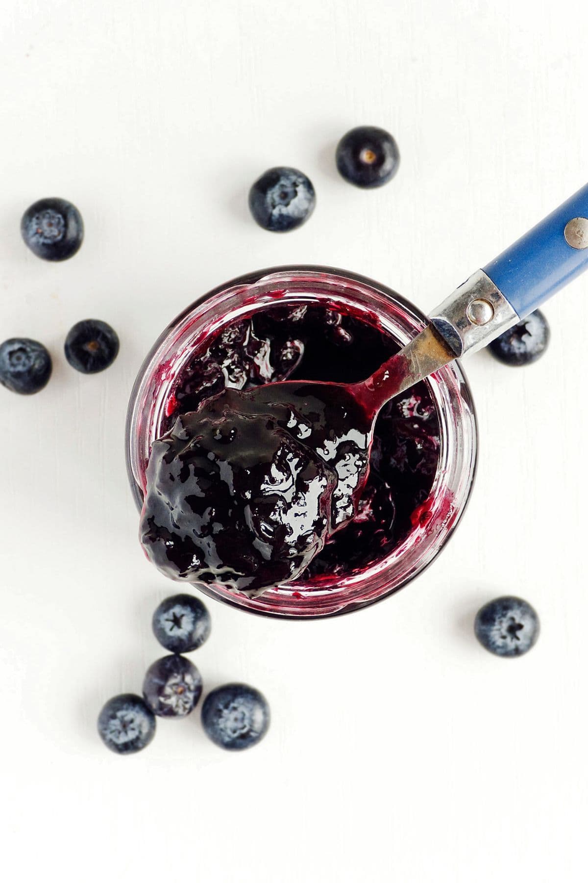 A jar of Blueberry Refrigerator Jam is open with a spoon dipped in it. Several whole blueberries are scattered around the jar on a white surface.