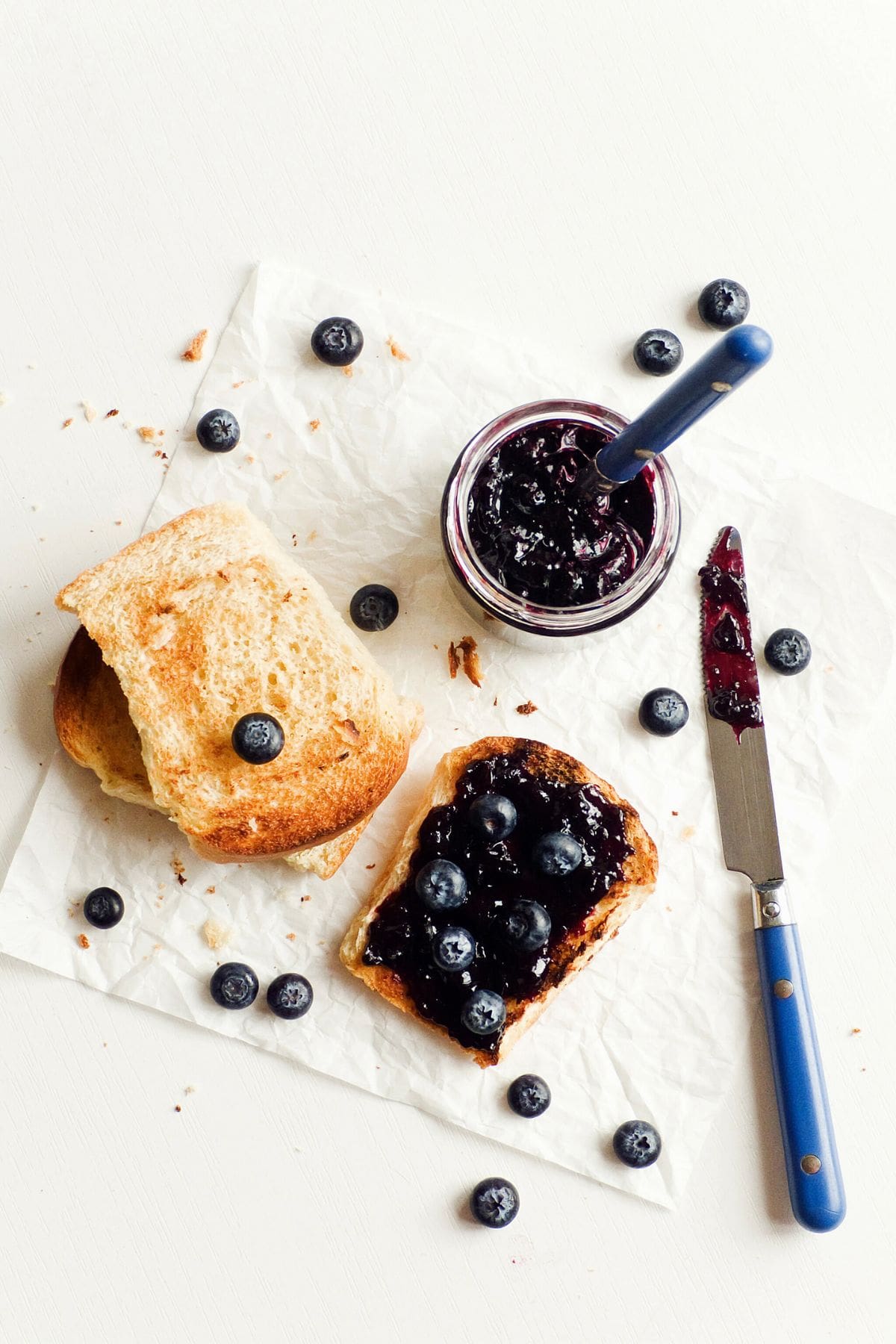 An image of a slice of toasted bread with 3-ingredient blueberry refrigerator jam and fresh blueberries on top. Another slice of toast is next to it, partially cut. A jar of blueberry jam with a spoon, and a knife with jam residue are placed nearby. Blueberries are scattered around.