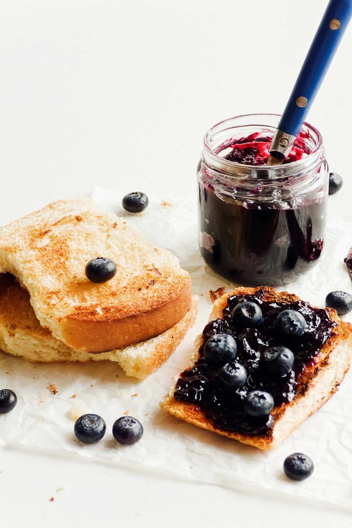 A slice of toast with 3-Ingredient Blueberry Refrigerator Jam and whole blueberries sits next to a partially eaten slice of plain toast. A jar filled with blueberry jam and a spoon is next to the toasts. Several whole blueberries are scattered around on a white surface.