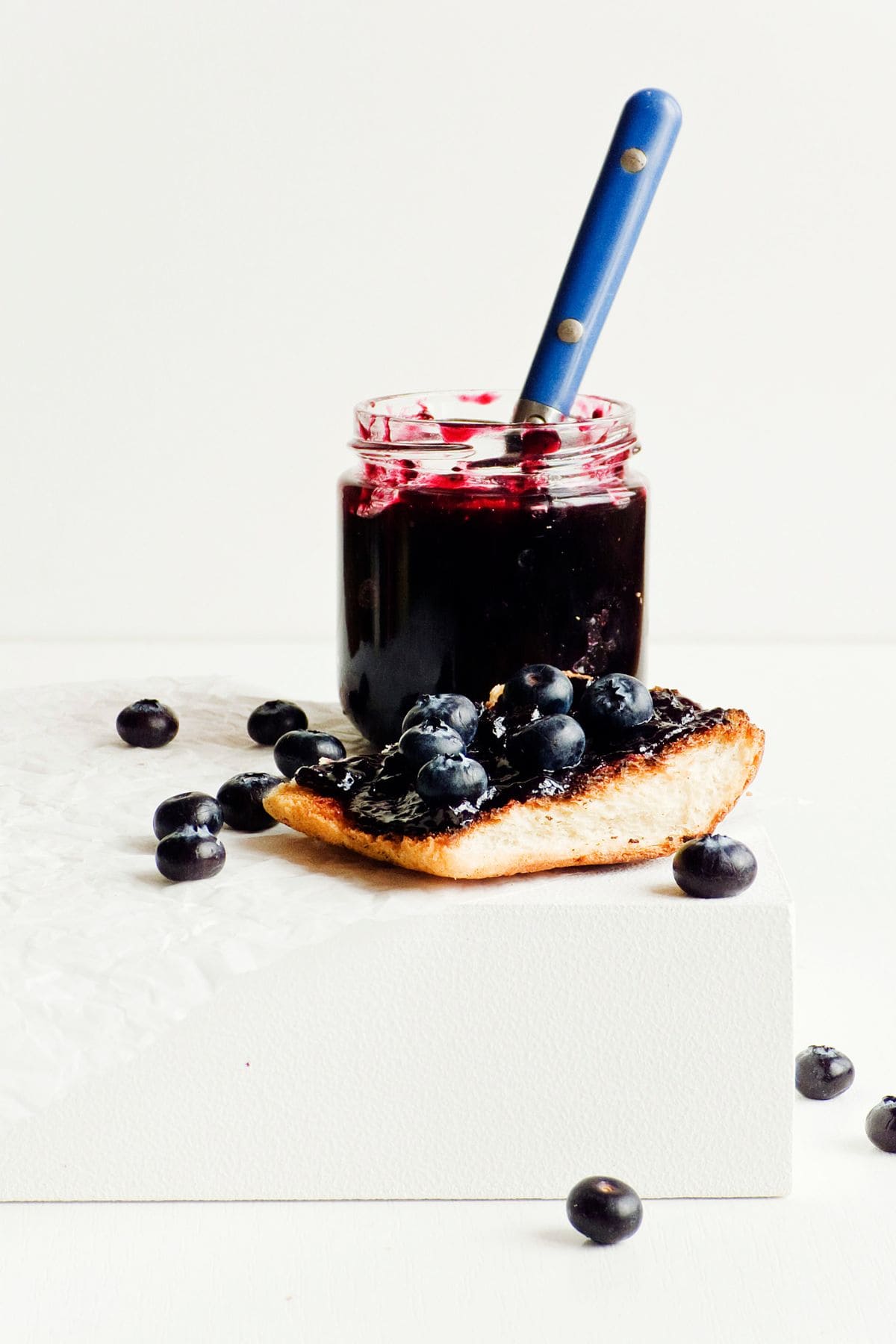 A jar of 3-ingredient blueberry jam with a blue-handled spoon placed inside it sits on a white surface. Next to the jar is a slice of bread spread with blueberry jam and topped with fresh blueberries. Several loose blueberries are scattered around the jar and bread, showcasing this refrigerator jam delight.