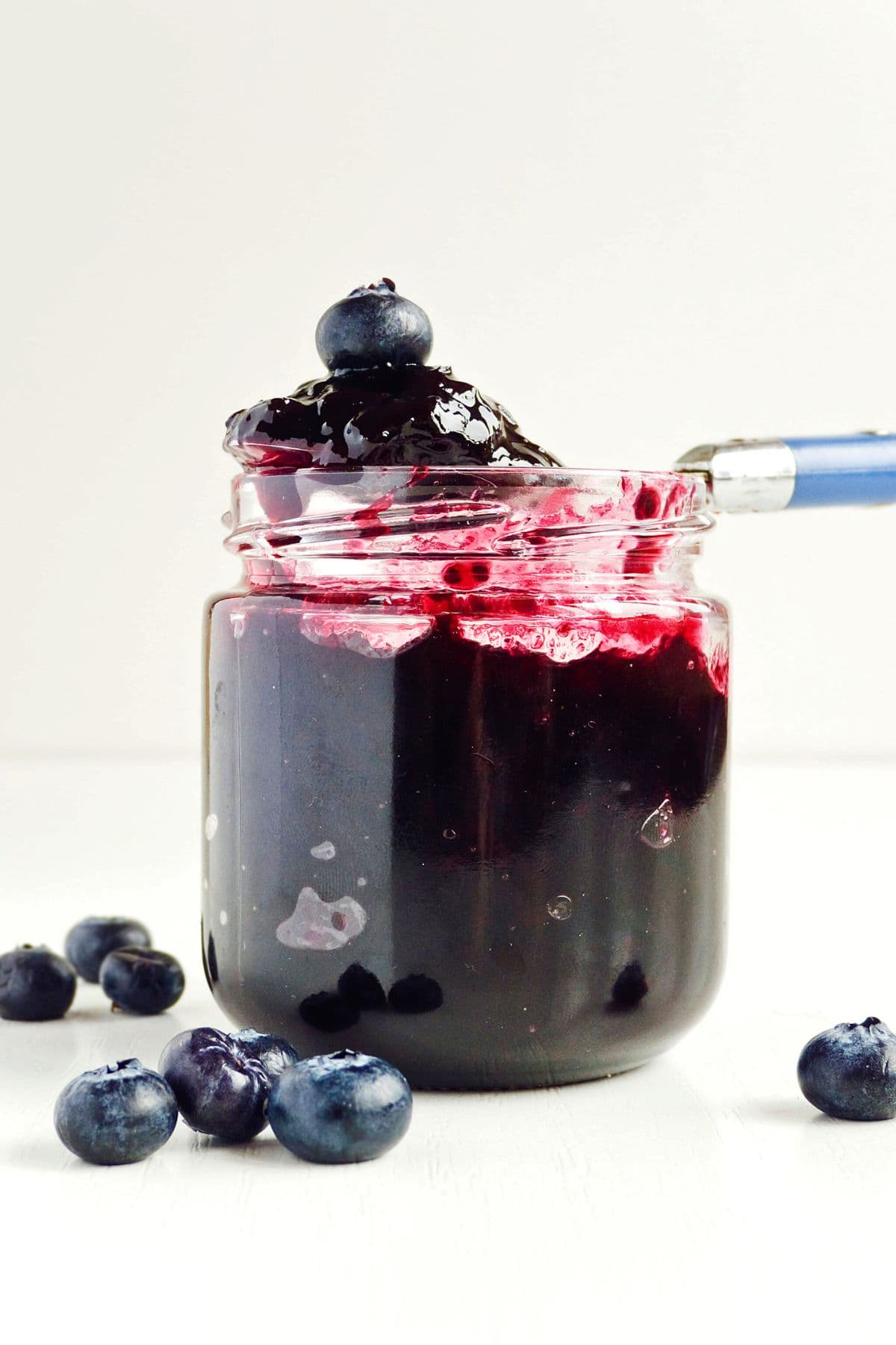 A clear jar filled with dark blueberry preserve, also known as 3-Ingredient Blueberry Jam, is displayed against a plain background. A spoon is partially submerged in the preserve, with some jam and a blueberry on top. Several fresh blueberries are scattered around the jar.