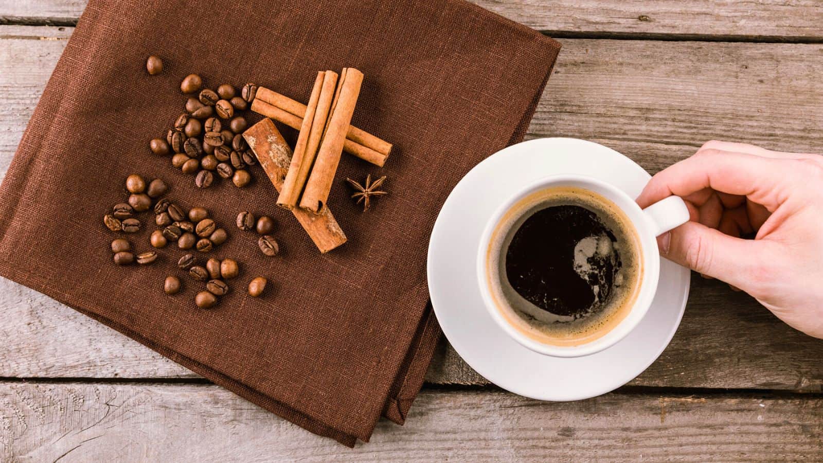 A hand holding a white cup filled with black coffee on a wooden table. Next to the cup, there is a brown cloth with coffee beans and cinnamon sticks scattered on it.