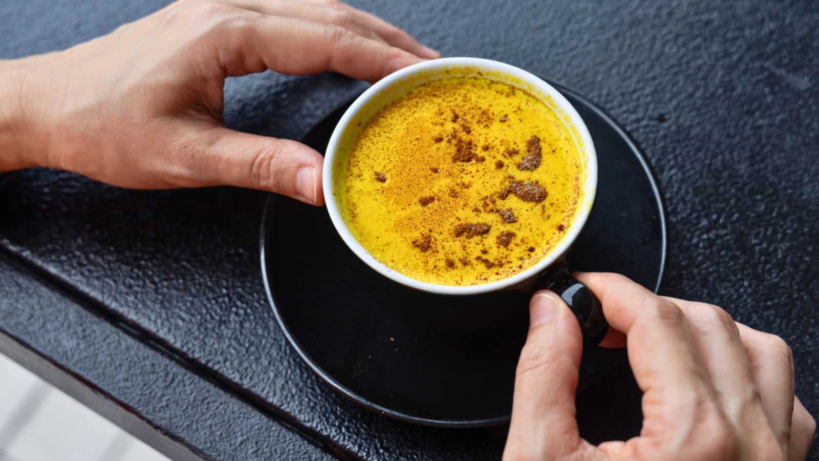 A person holds a cup of golden turmeric latte, sprinkled with spices, on a black saucer. The cup is on a black table. The person's hands are visible but not the rest of their body.