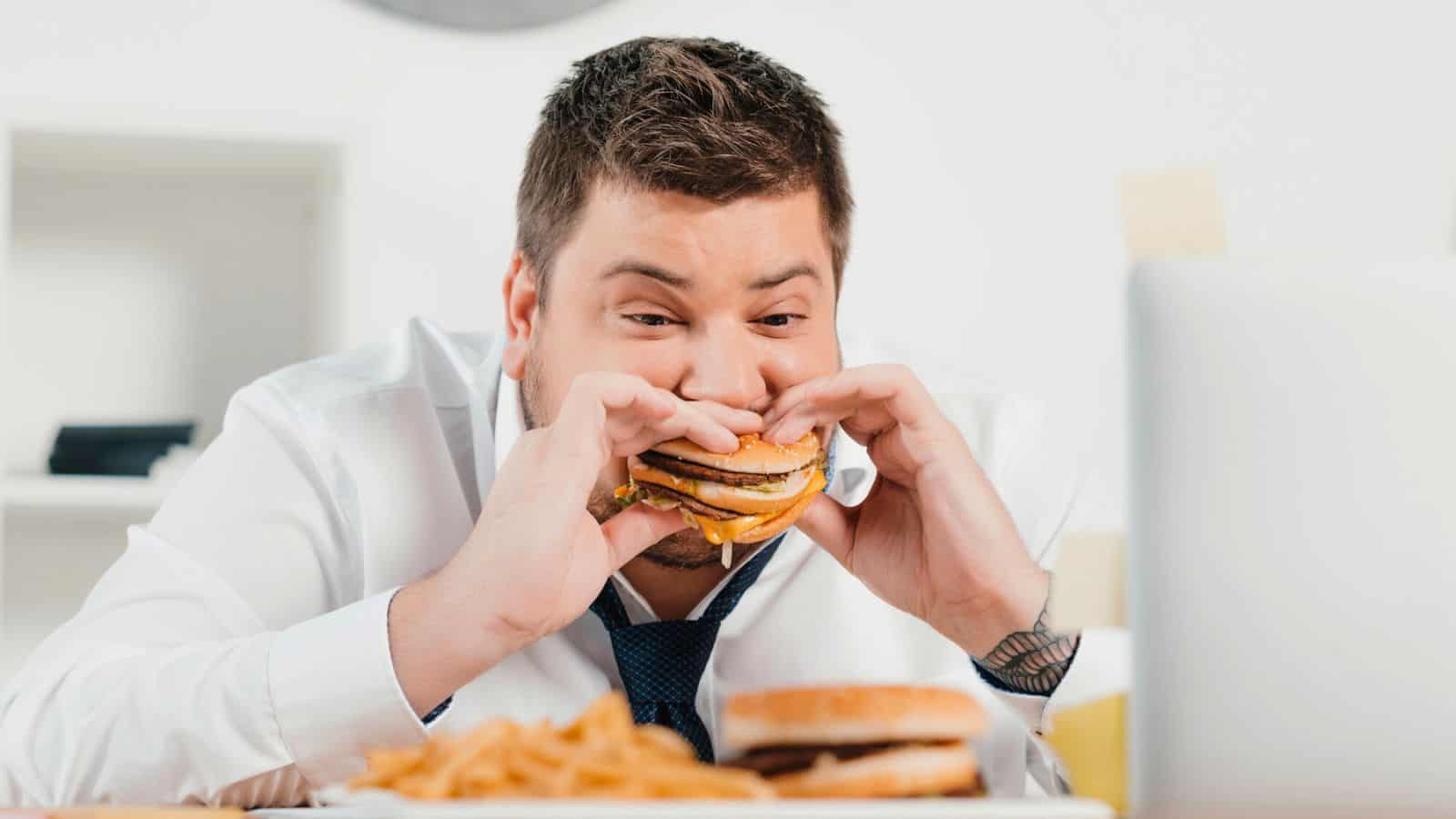 A man in a white dress shirt and blue tie is sitting at a table, intently looking at a laptop screen. He is eating a large burger with both hands. In front of him are fries and another burger. The setting appears to be an office.