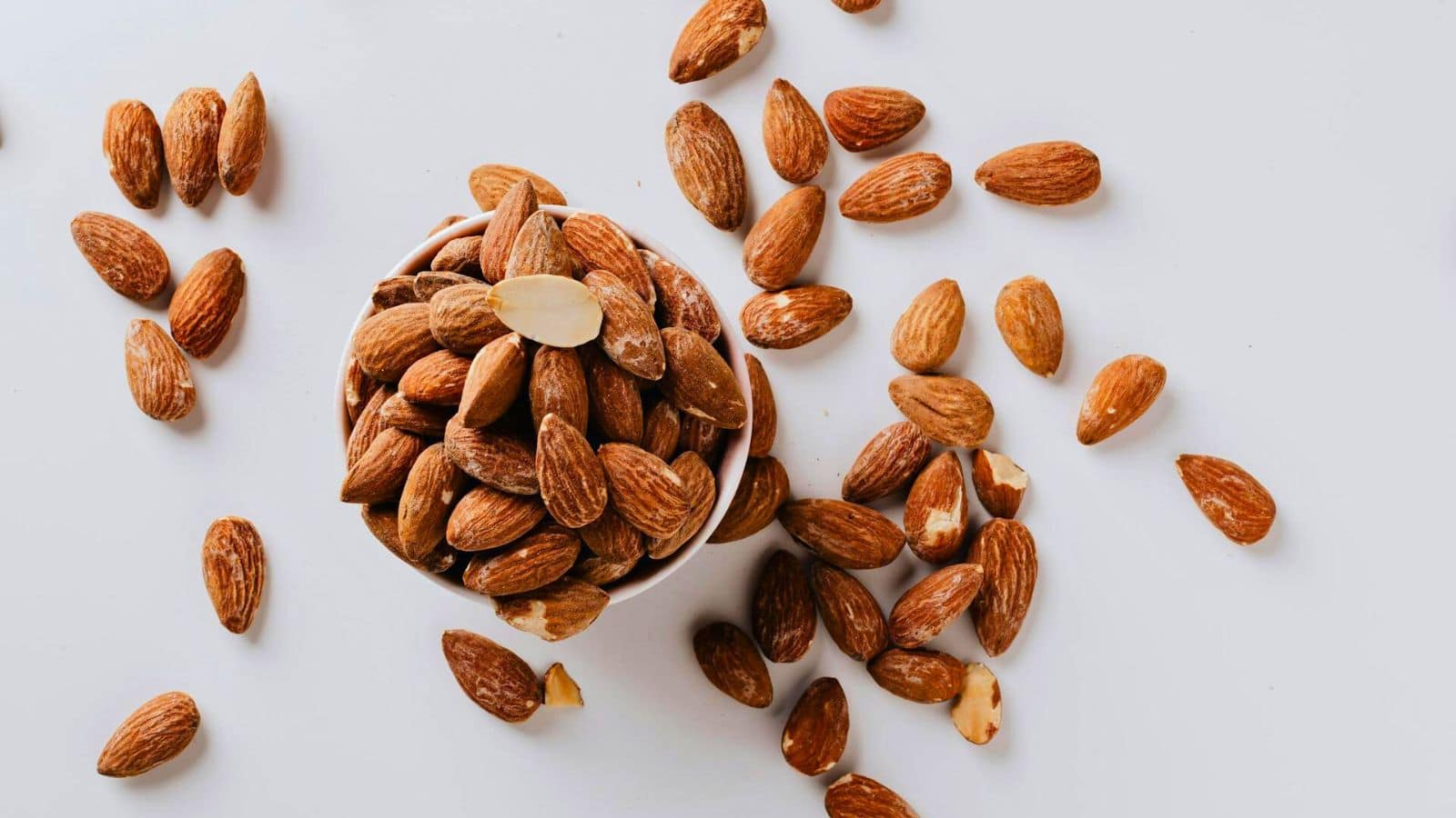 A small white bowl filled with whole almonds is placed on a white surface. Several almonds are scattered around the bowl, creating a spread-out arrangement. One almond at the top of the bowl is slightly chipped.