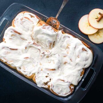 A glass baking dish filled with frosted cinnamon rolls, accompanied by a spoon. Beside the dish are two apple slices and three cinnamon sticks arranged on a dark surface, perfect for those seeking apple-icious recipes for easy weeknight meals.