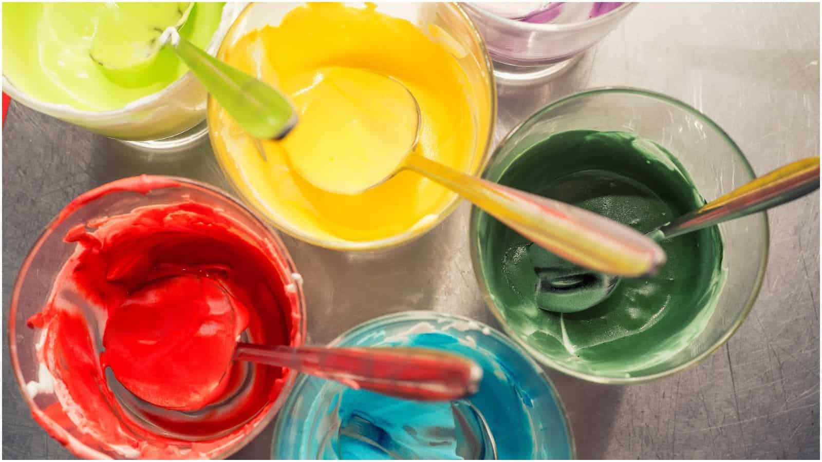 Various glass bowls of brightly colored icing in green, yellow, red, blue, and purple with matching spoons are arranged on a reflective surface. Each bowl is filled with a different hue, creating a vibrant display.