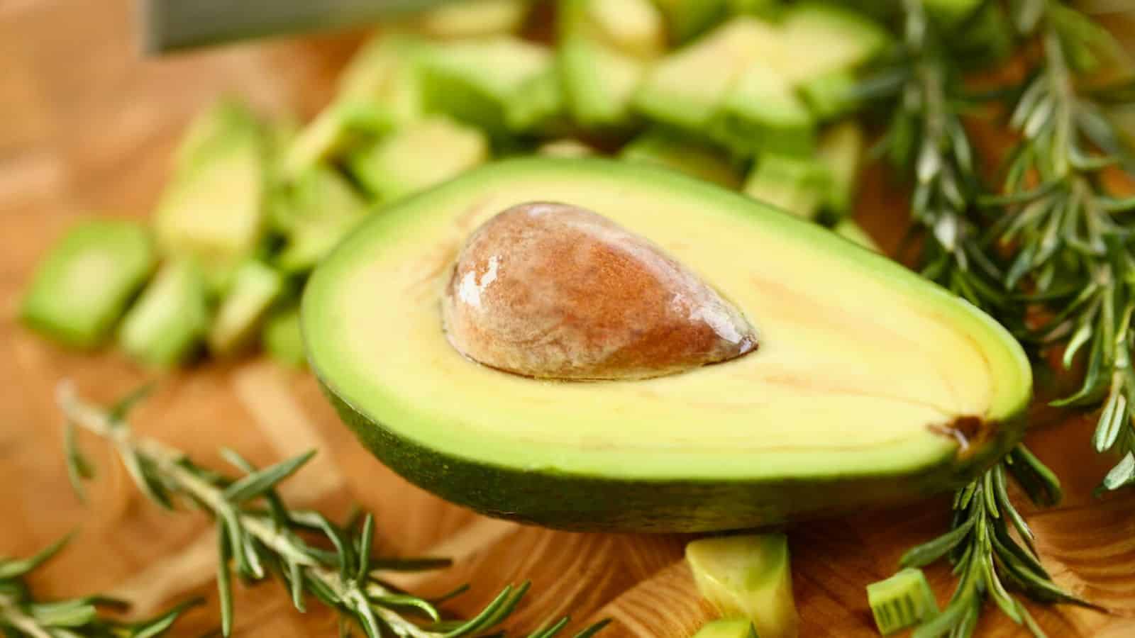 A halved avocado with the seed still inside is placed on a wooden cutting board. Surrounding it are diced pieces of avocado and some sprigs of rosemary. The background is slightly blurred, with part of a knife visible at the top.