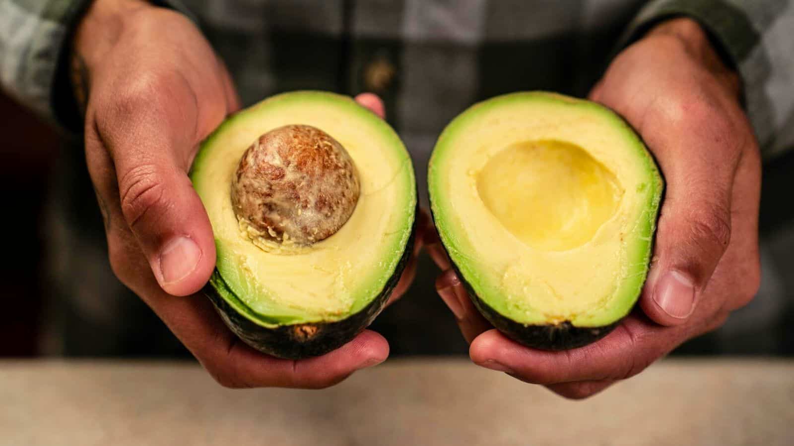 A person holds a halved avocado with both hands. The left half shows an avocado pit, while the right half has the pit removed, revealing smooth, green flesh. The person is wearing a plaid shirt. The background is blurry and indistinct.