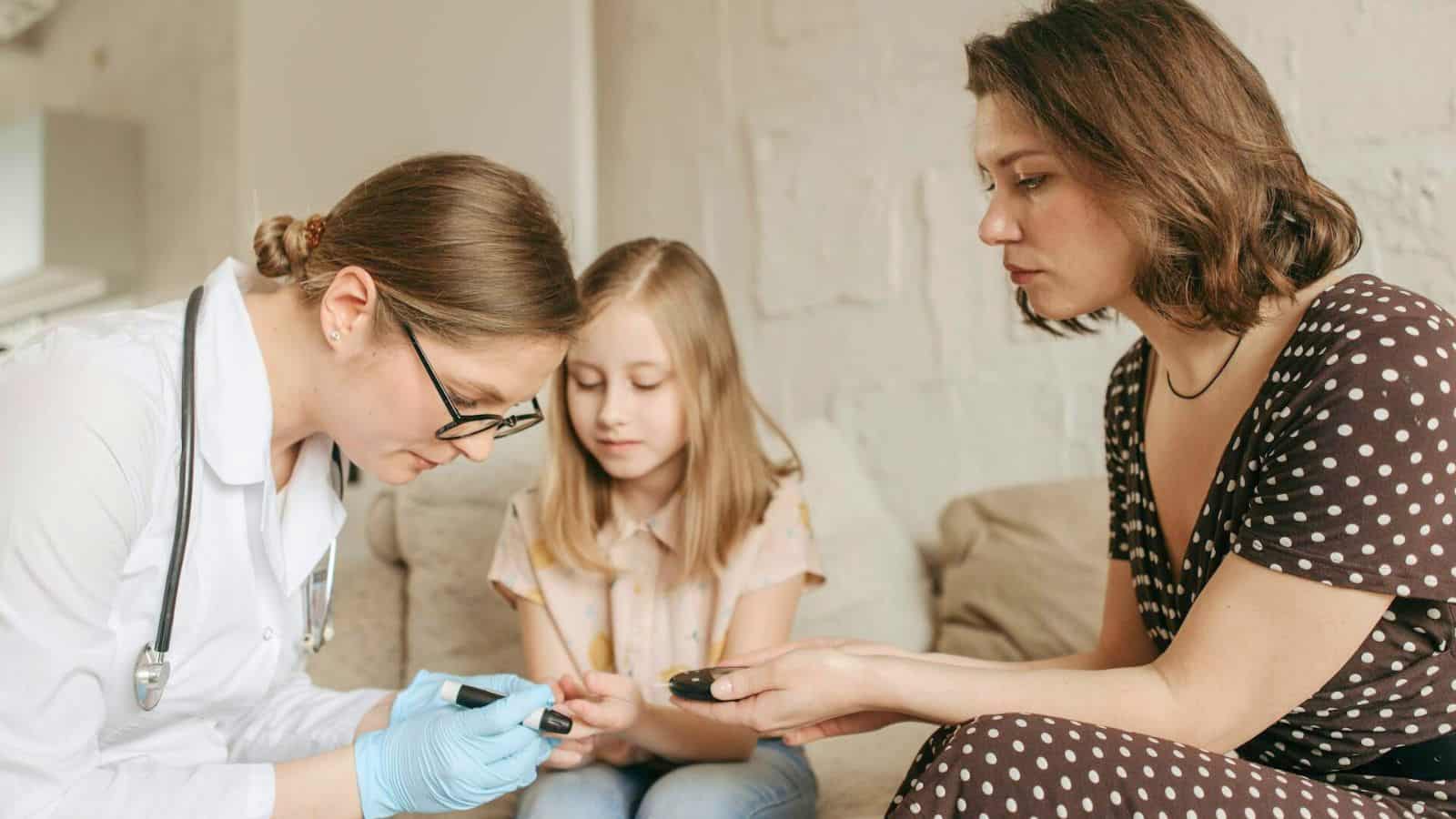 A female doctor with glasses and blue gloves measures blood sugar using a glucometer on a woman’s hand while a young girl watches. They are sitting on a beige couch beside a white wall.