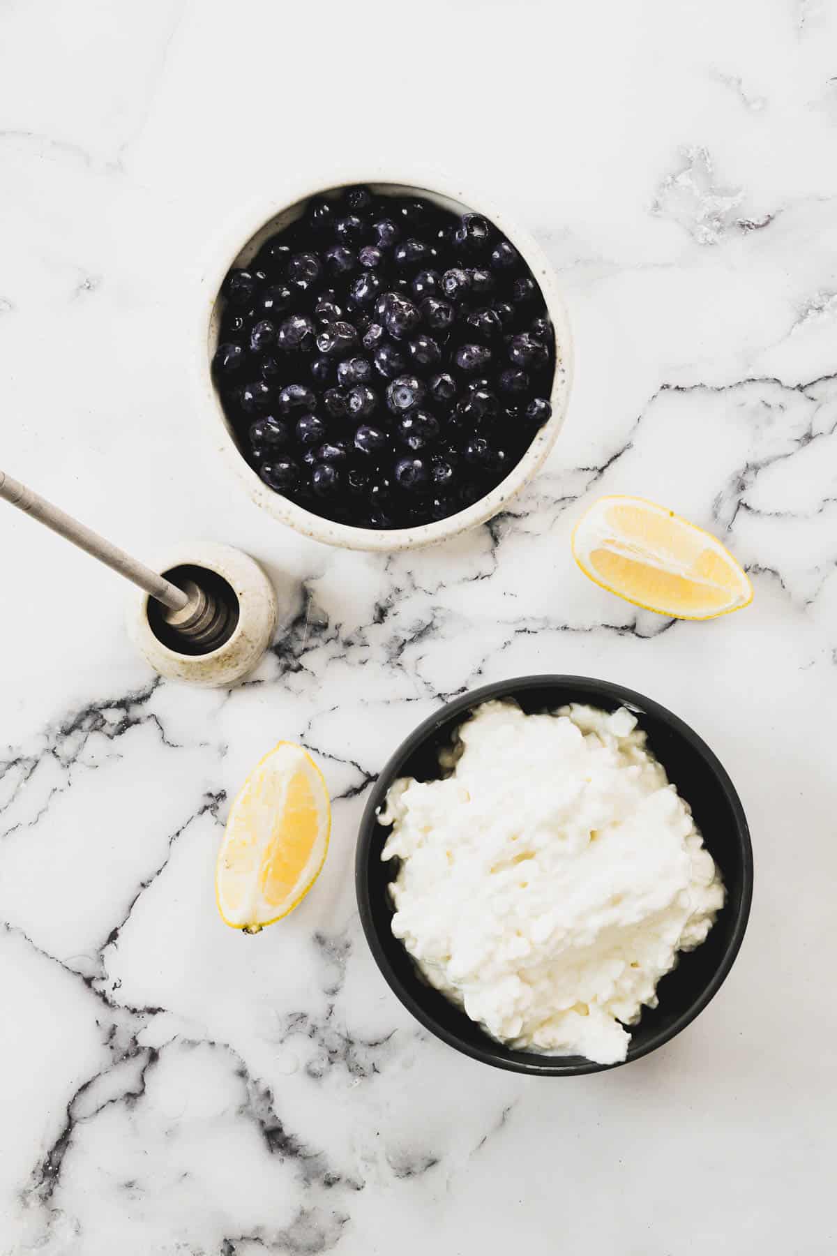 A top view of a bowl of blueberries and a bowl of cottage cheese on a marble surface. Beside them are a honey dipper with honey and two lemon wedges.