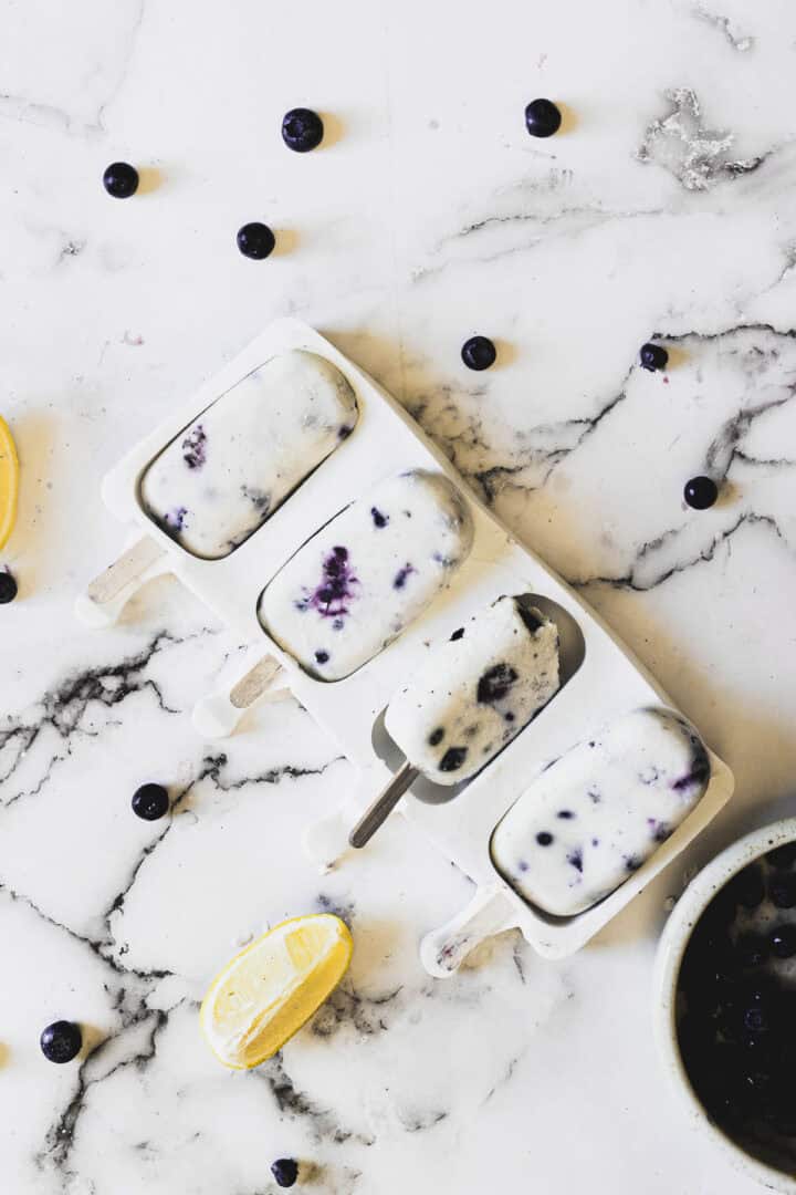 Four blueberry and cream popsicles are arranged in a white mold on a marble surface. There are scattered blueberries and a lemon wedge around the mold. One popsicle is partially melted with a bite taken out of it, resting on the mold.