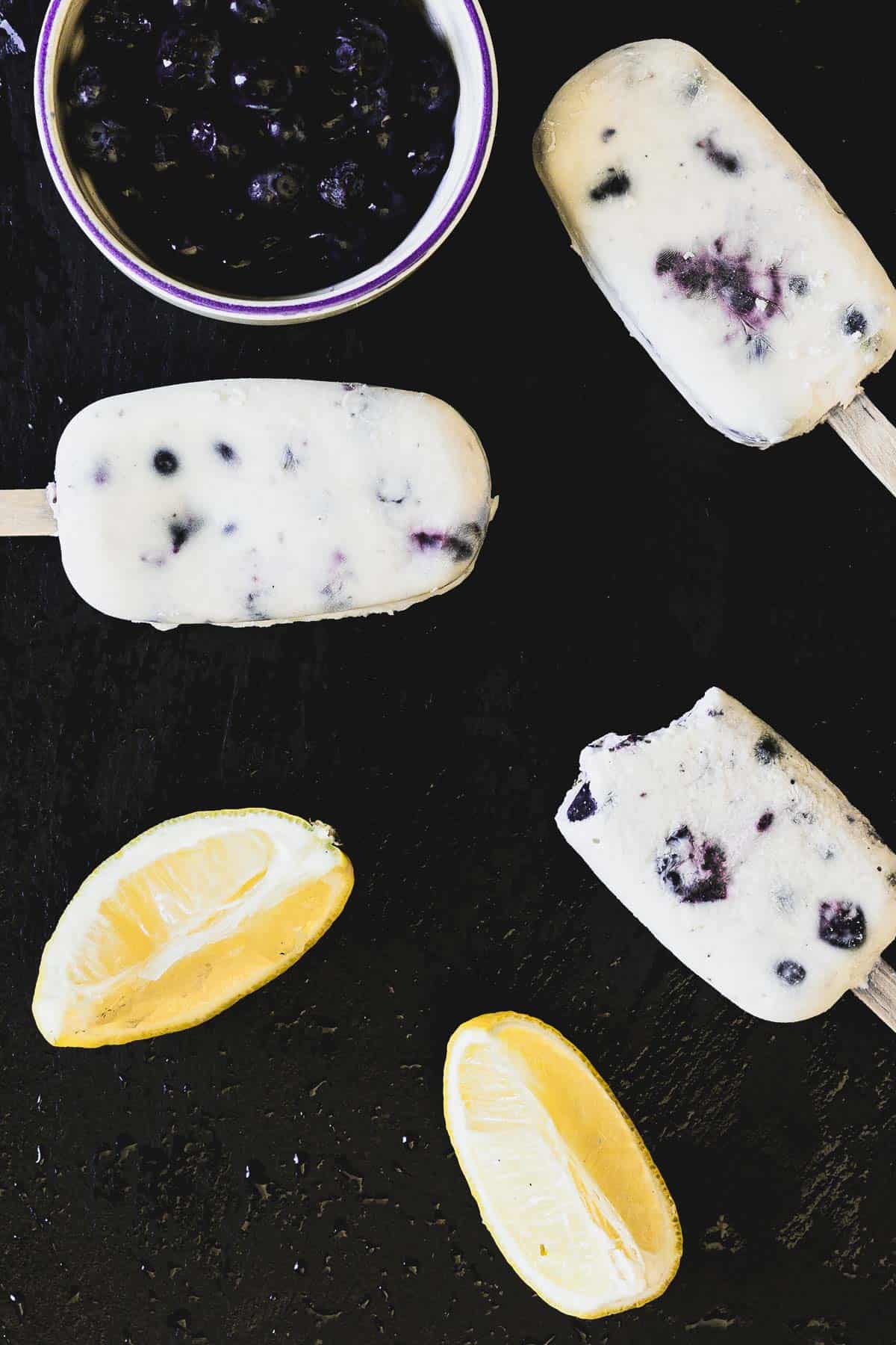 A top view of three blueberry yogurt popsicles and two lemon wedges on a black surface. One popsicle has a bite taken out of it. A small bowl of blueberries is placed nearby.