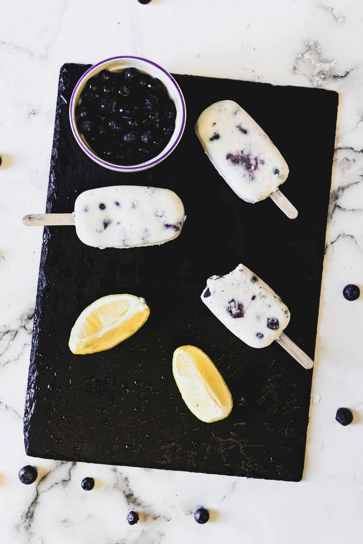 Three blueberry yogurt popsicles and two lemon wedges are displayed on a black slate board. A small bowl filled with blueberries is placed at the top of the slate board. The background is a white marble surface with scattered blueberries.