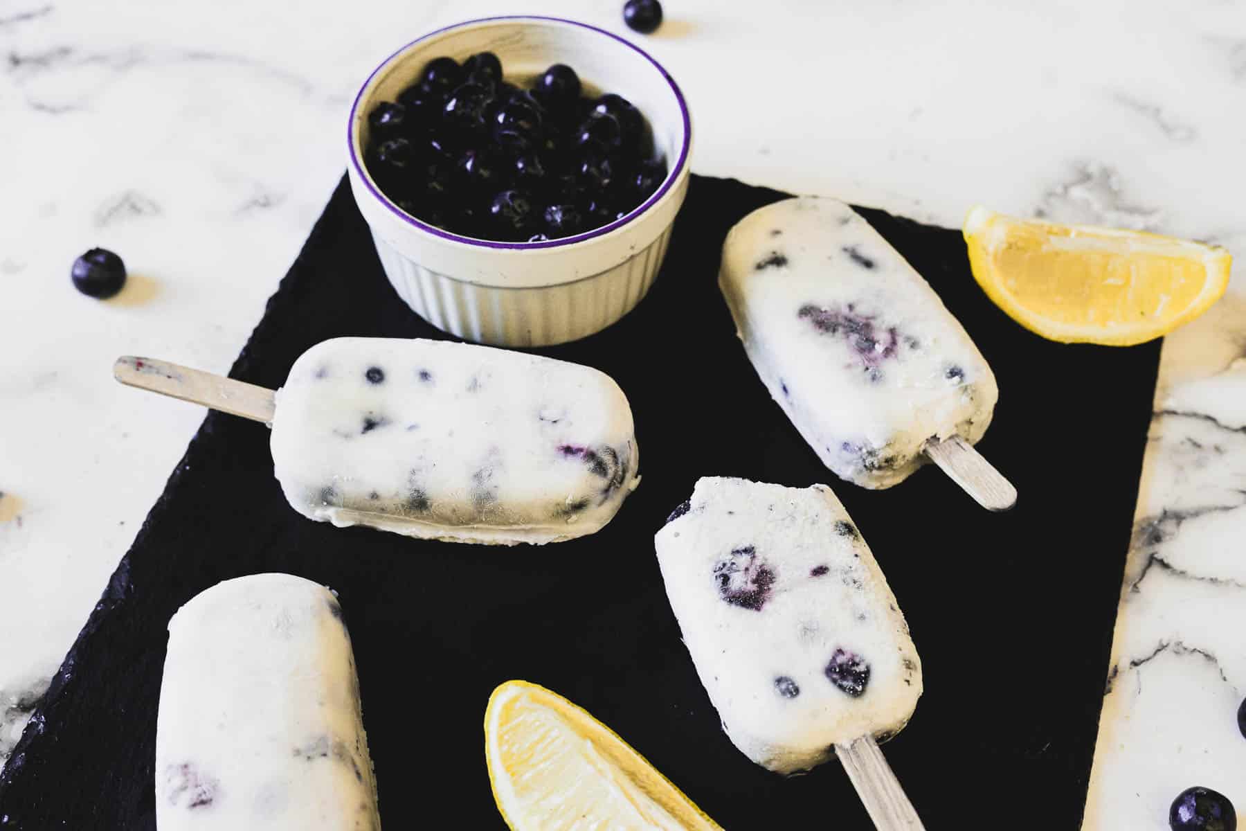 A slate platter holds five blueberry yogurt popsicles on wooden sticks. A small bowl containing fresh blueberries and a couple of lemon wedges are also on the platter. The background features a white marble surface.