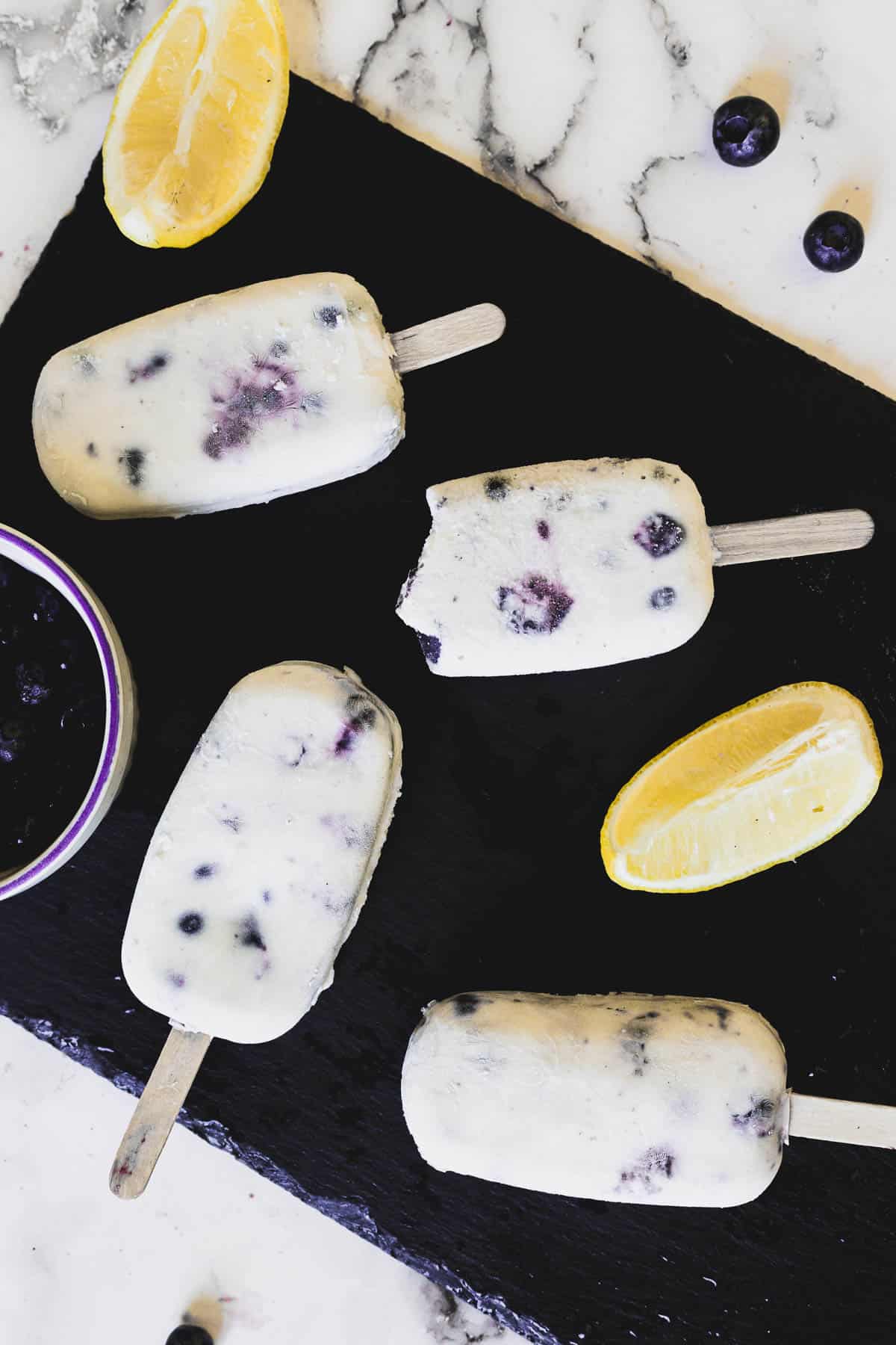 Five blueberry popsicles are positioned on a black slate, with one partially eaten. Two lemon wedges and a small bowl of blueberries accompany the popsicles, all set on a white marble surface.