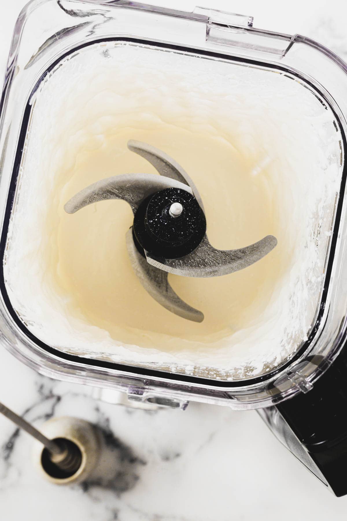 Image of a blender with a transparent container filled with a creamy, light-colored mixture. The sharp, four-blade assembly is visible at the center. The blender sits on a white marble countertop, and a black utensil is partially seen in the lower left corner.