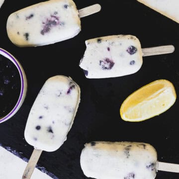 Four blueberry ice cream popsicles on sticks are arranged on a dark surface. One popsicle has a bite taken out of it. A bowl containing blueberries and a wedge of lemon are positioned nearby.