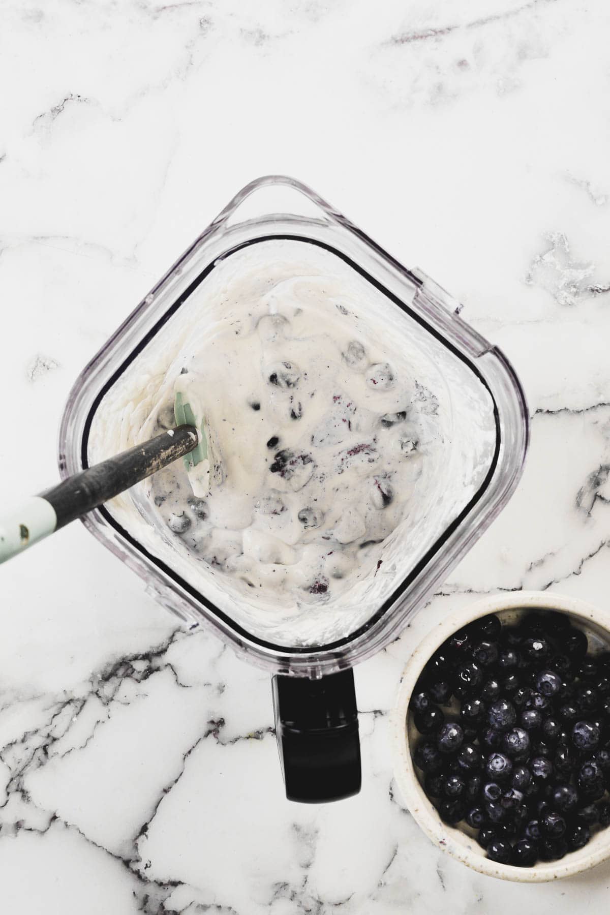 Overhead view of a blender filled with a mixture of yogurt and blueberries with a spatula inside. Next to the blender is a small bowl filled with additional blueberries. The background is a marble countertop.