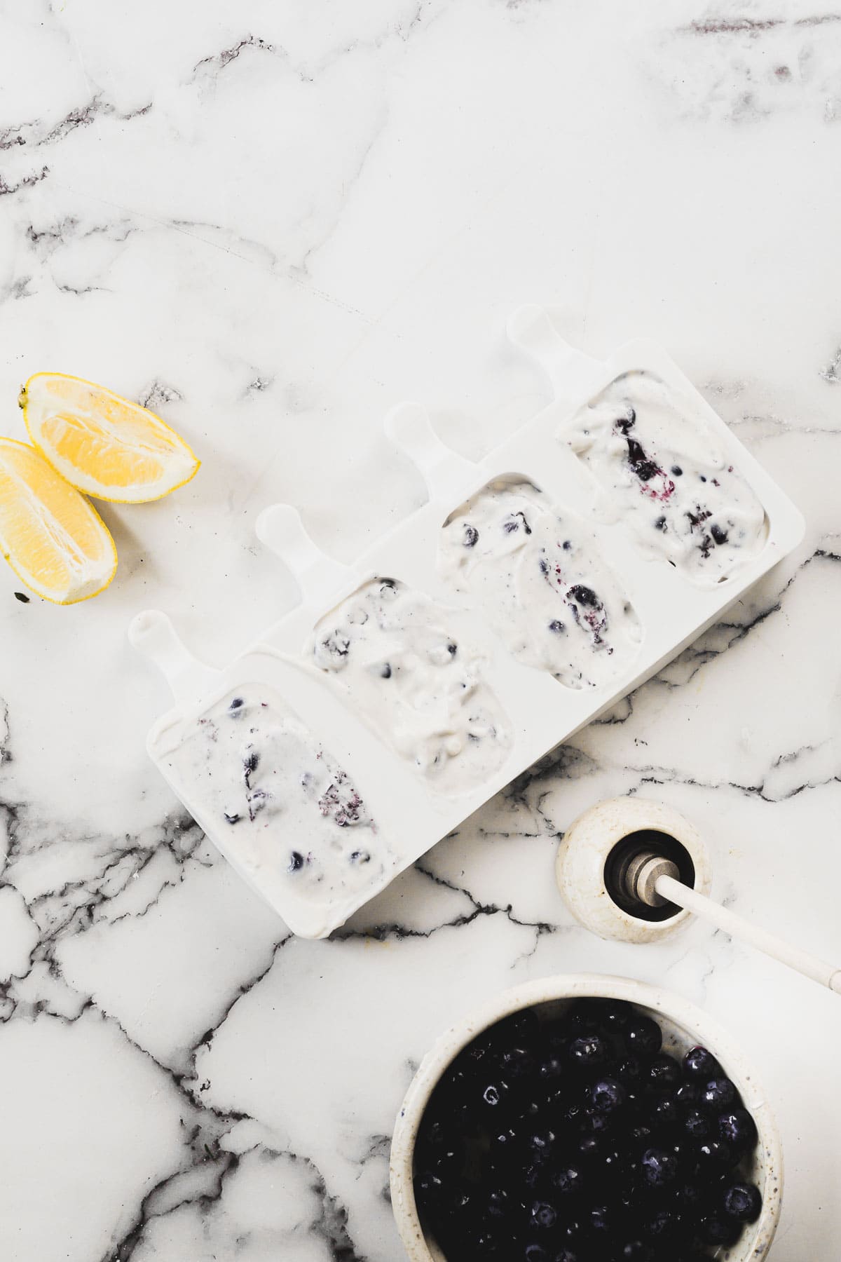 A set of four homemade blueberry yogurt popsicles in a white mold is placed on a marble countertop. Next to the mold are two lemon wedges and a white bowl containing fresh blueberries. A portion of the countertop features a milk spill.