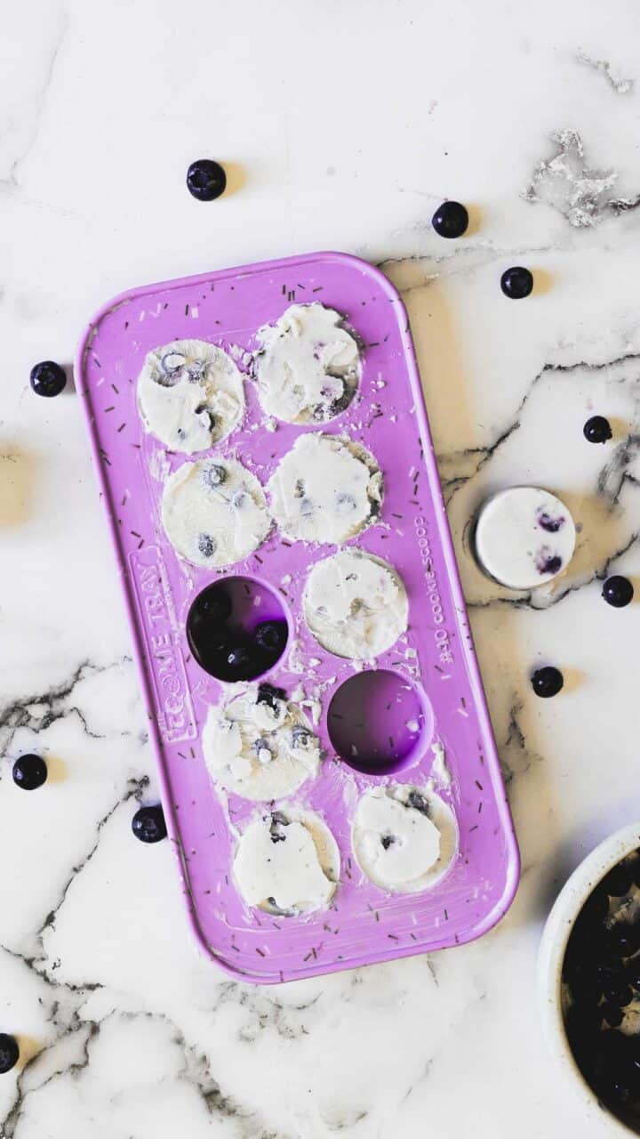 A purple silicone mold holds eight frozen yogurt bites, each embedded with blueberries. One bite has been removed and is placed to the right of the mold. The mold and bites are on a white marble surface scattered with more blueberries.