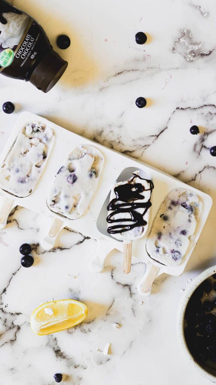 Four blueberry and yogurt popsicles are in a mold on a marbled counter, with one popsicle being held up covered in chocolate syrup. There are scattered blueberries, a lemon wedge, and a bottle of chocolate syrup nearby.