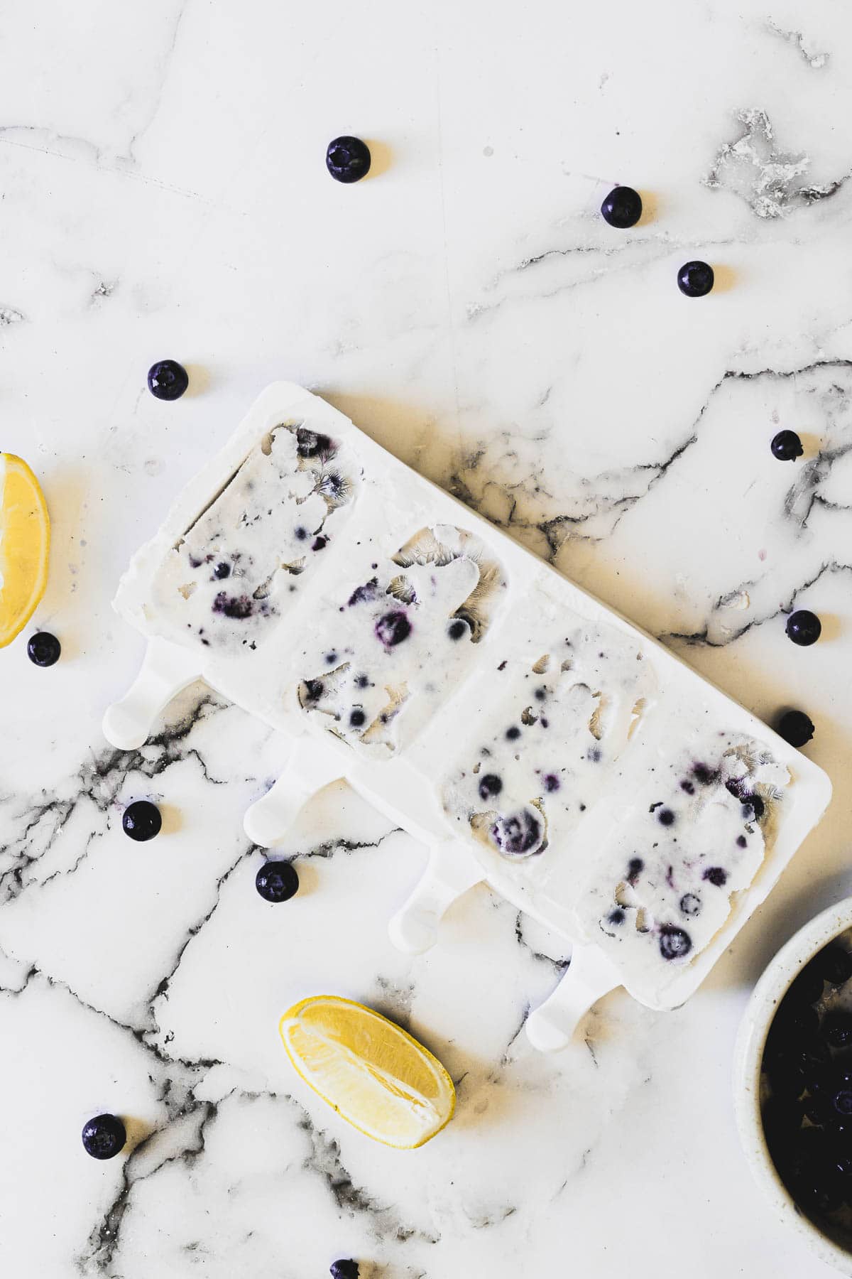 A white popsicle mold filled with blueberry yogurt popsicles sits on a marble countertop. Blueberries are scattered around the mold, along with two lemon wedges and a bowl partially visible at the bottom right corner.