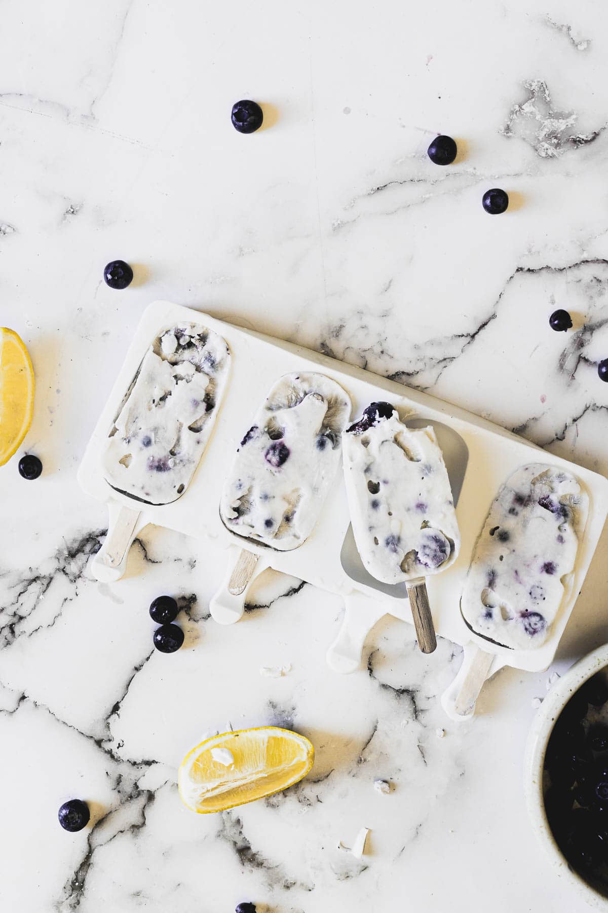 Four blueberry and cream popsicles on a white marble surface with scattered blueberries and lemon wedges. One popsicle is partially eaten and the mold is visible beneath. A bowl with contents is partially visible in the bottom right corner.