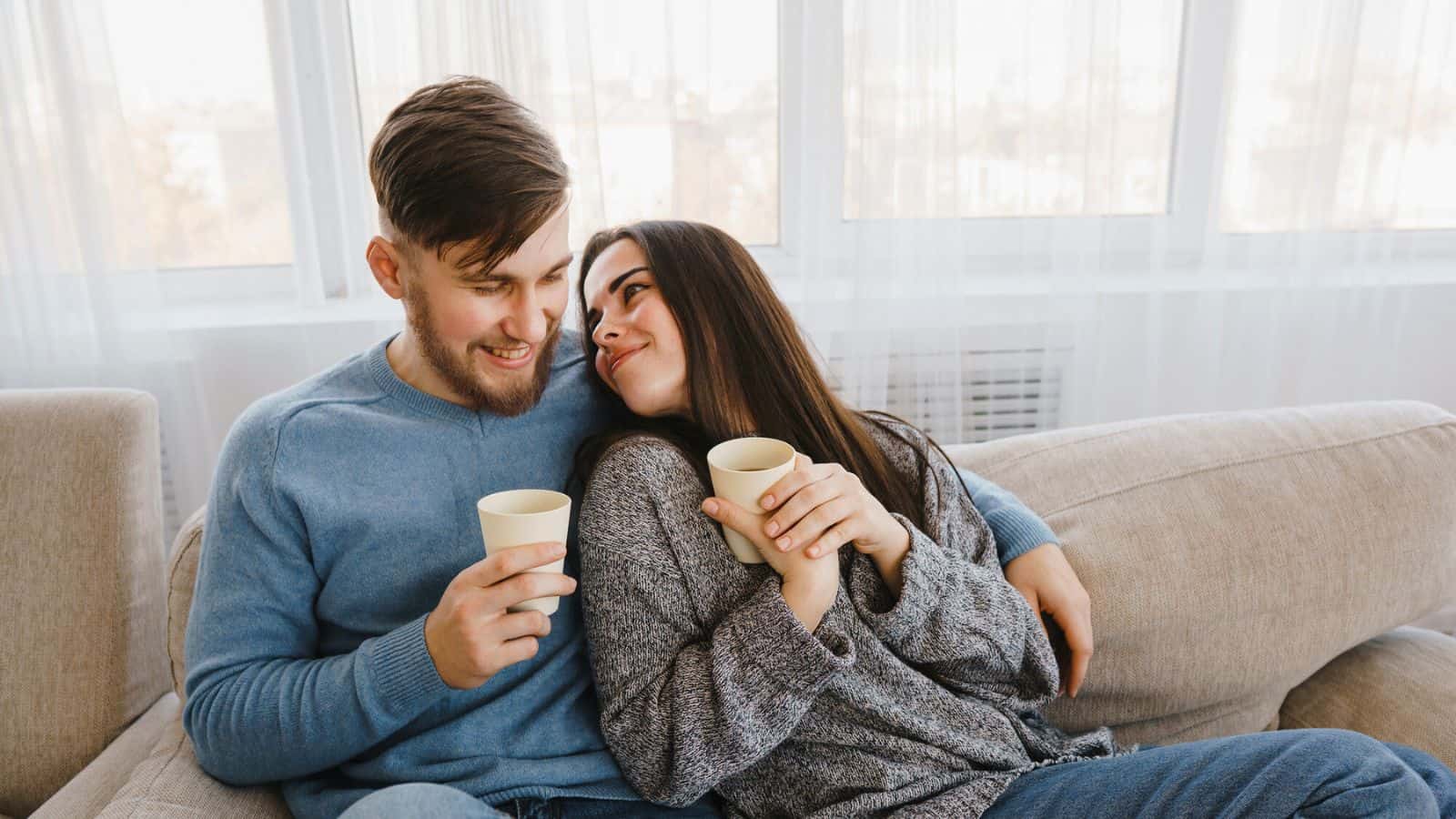 A man in a blue sweater and a woman in a grey cardigan sit closely together on a beige couch, each holding a cup. They are looking at each other and smiling. A large window with sheer curtains is visible in the background.