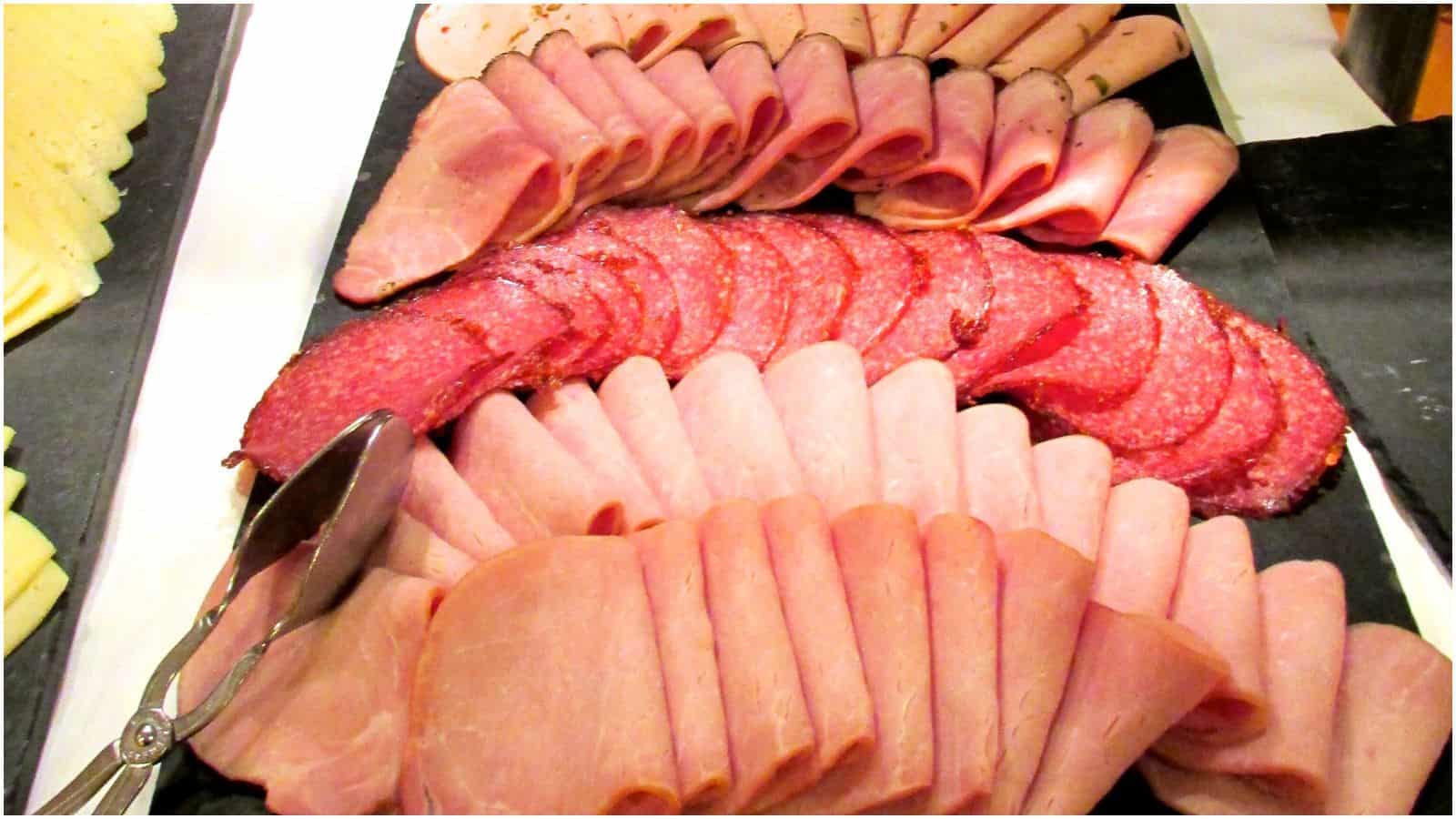 A platter displaying an assortment of sliced deli meats, including ham, turkey, and salami, arranged in overlapping layers. A pair of metal tongs rests on the left side of the platter.