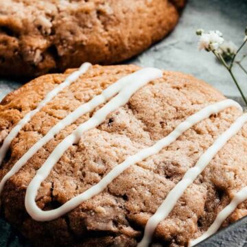 A close-up of a freshly baked Good-For-You Cookie with white icing drizzled on top. The cookie, reminiscent of Childhood Favorites, rests on parchment paper with small white flowers placed nearby for decoration.