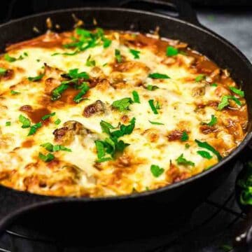 A skillet filled with a cheesy, baked low-carb delight garnished with green herbs. Shredded cheese and fresh herbs are in small bowls beside the skillet. The dish, perfect for low-carb dinners, is placed on a round cooling rack over a dark surface.