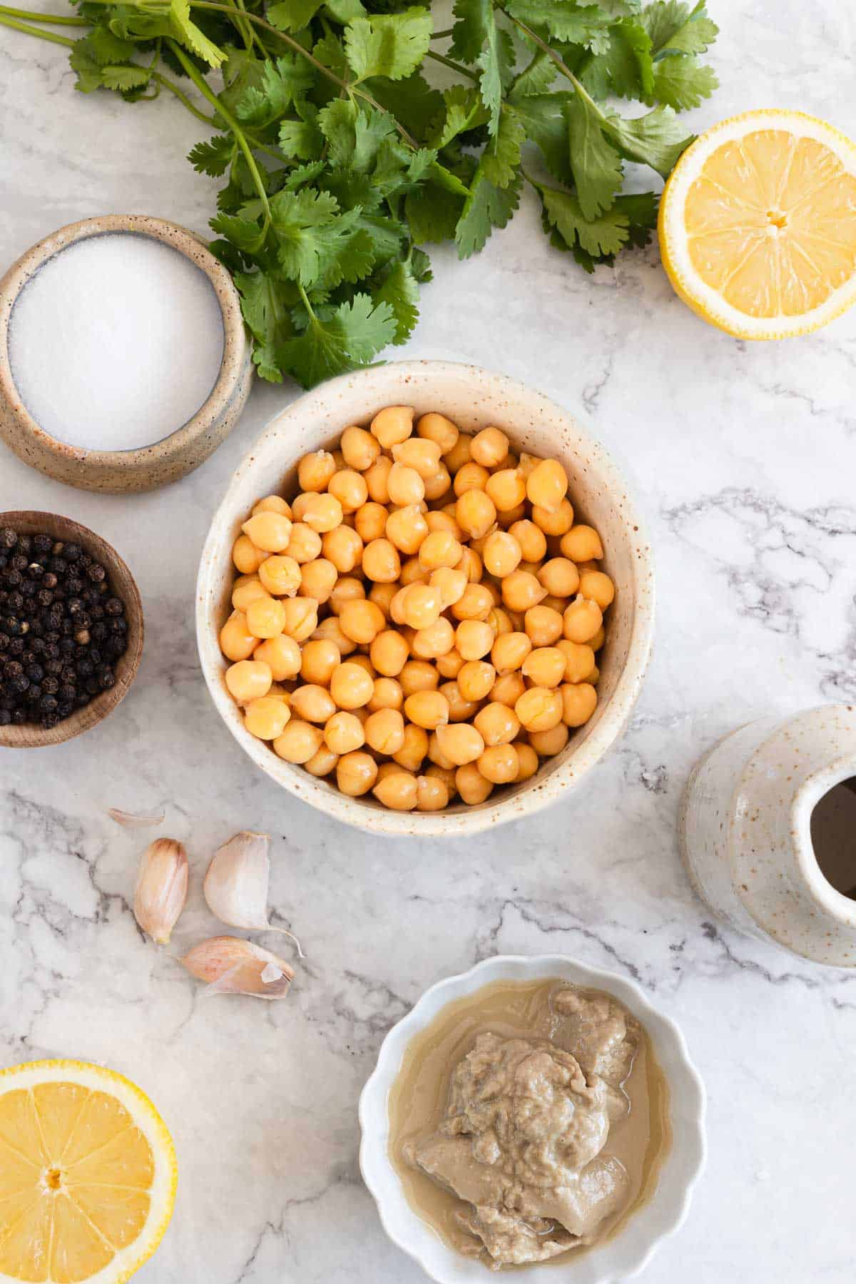 A marble countertop with ingredients for making hummus. A bowl of chickpeas is at the center, surrounded by halved lemons, garlic cloves, black peppercorns, salt, fresh cilantro, and a small dish of tahini. A small pitcher is also present.
