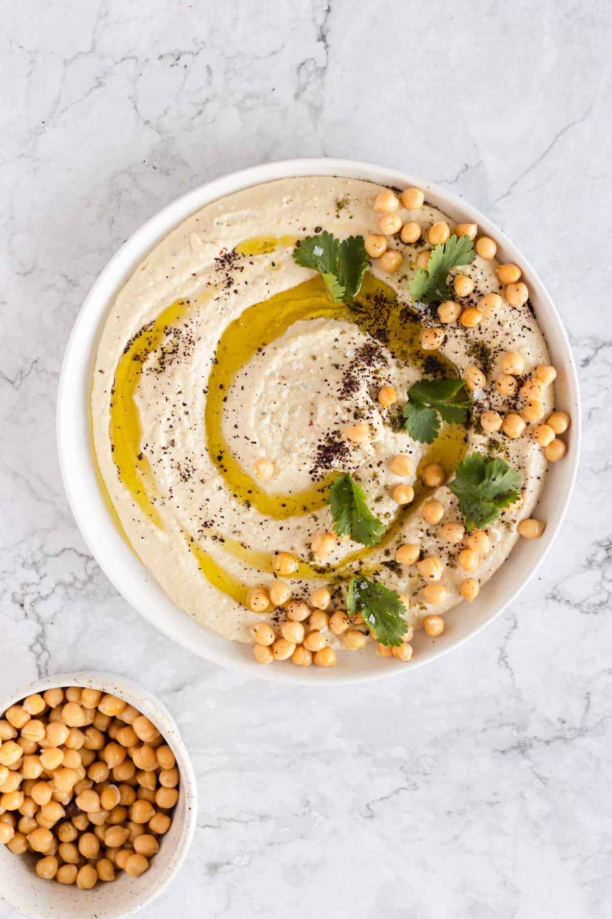 A bowl of hummus topped with a swirl of olive oil, chickpeas, cilantro, and pepper, sits on a marble surface. A small bowl of chickpeas is next to it.
