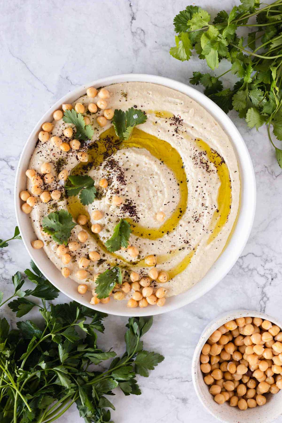 A bowl of hummus is garnished with chickpeas, olive oil, black pepper, and cilantro. Fresh cilantro and parsley are placed around the bowl, with a smaller bowl of chickpeas to the right. The background is a marbled white surface.