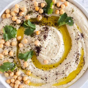 A bowl of canned chickpea hummus garnished with whole chickpeas, parsley leaves, a drizzle of olive oil, and a sprinkle of ground spices. The hummus is presented in a white bowl placed on a light grey marble surface.