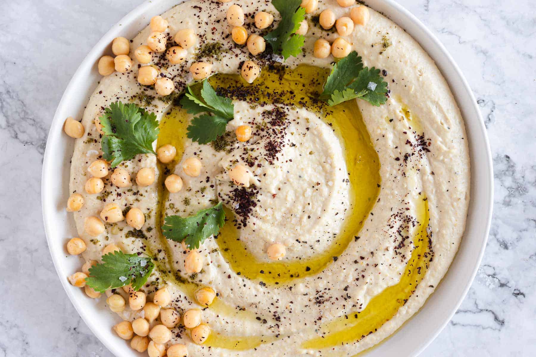 A bowl of hummus garnished with whole chickpeas, cilantro leaves, a drizzle of olive oil, and a sprinkle of spices. The hummus has a smooth texture and is presented in a round, white bowl placed on a marble surface.
