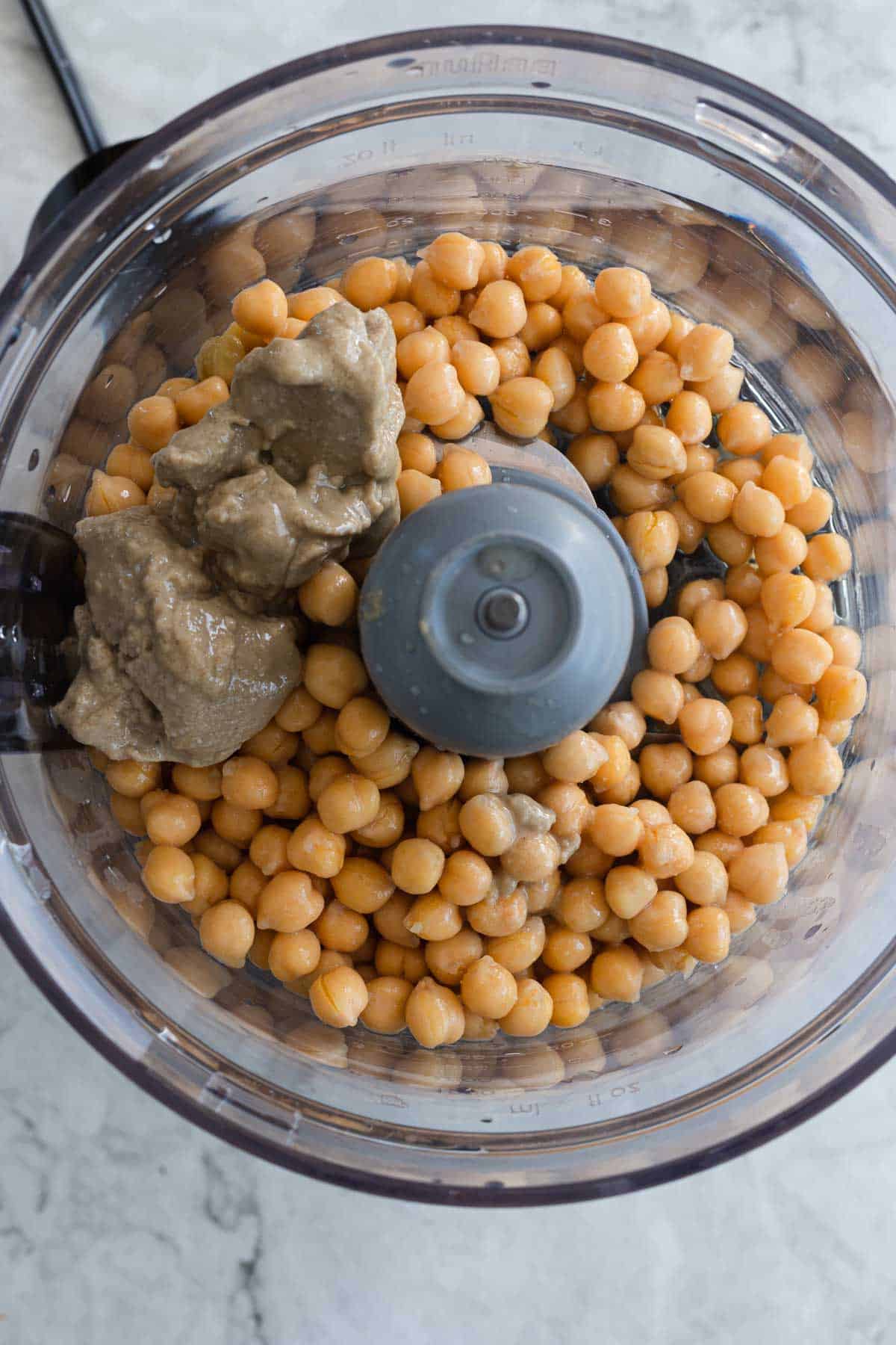 A food processor bowl contains chickpeas and two blobs of tahini.
