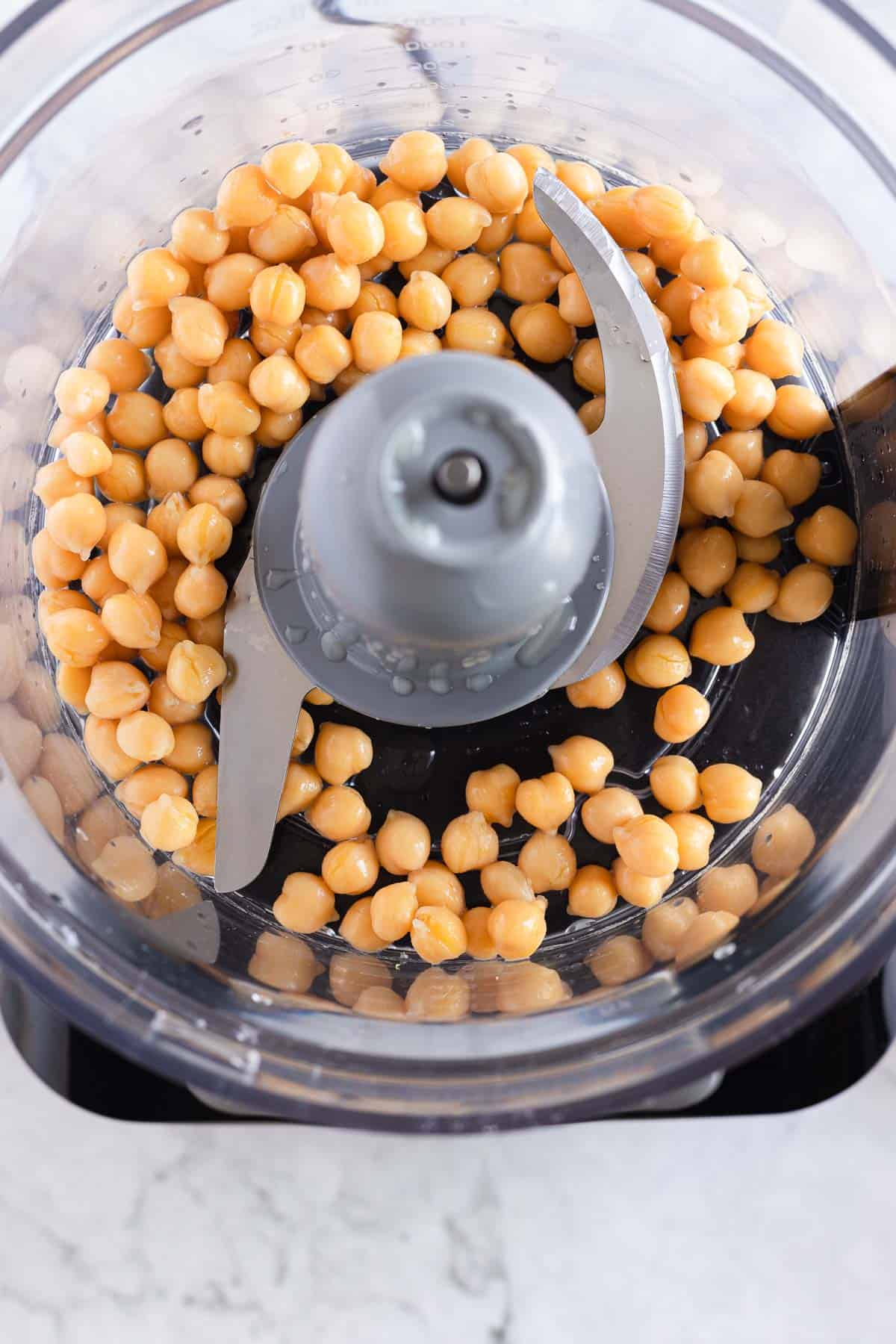 A food processor bowl filled with chickpeas, ready to be processed. The stainless steel blade is visible in the center of the bowl. The food processor is placed on a white marble surface.