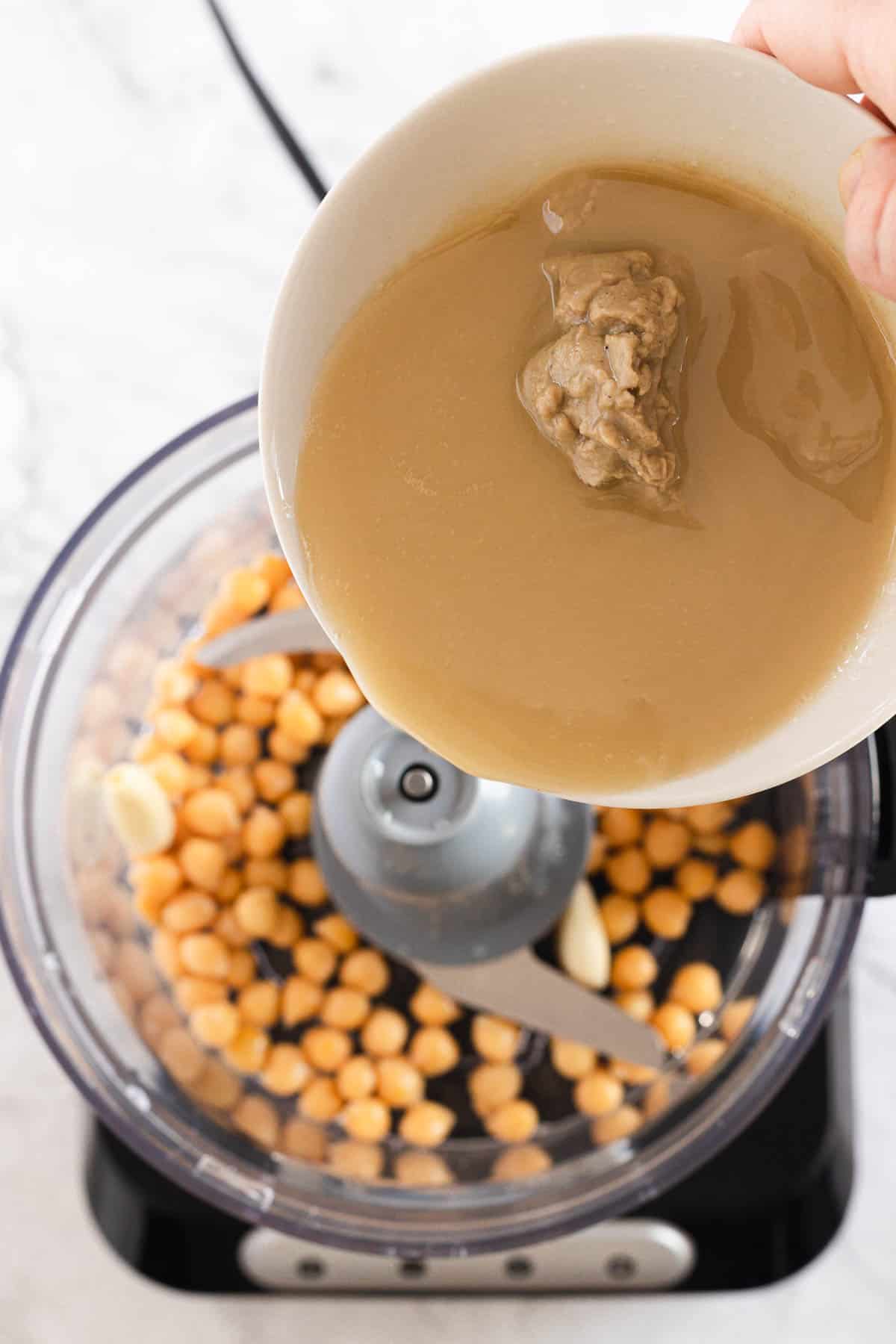 A hand is pouring tahini from a white bowl into a food processor containing chickpeas and garlic cloves. The food processor is placed on a white marble countertop.