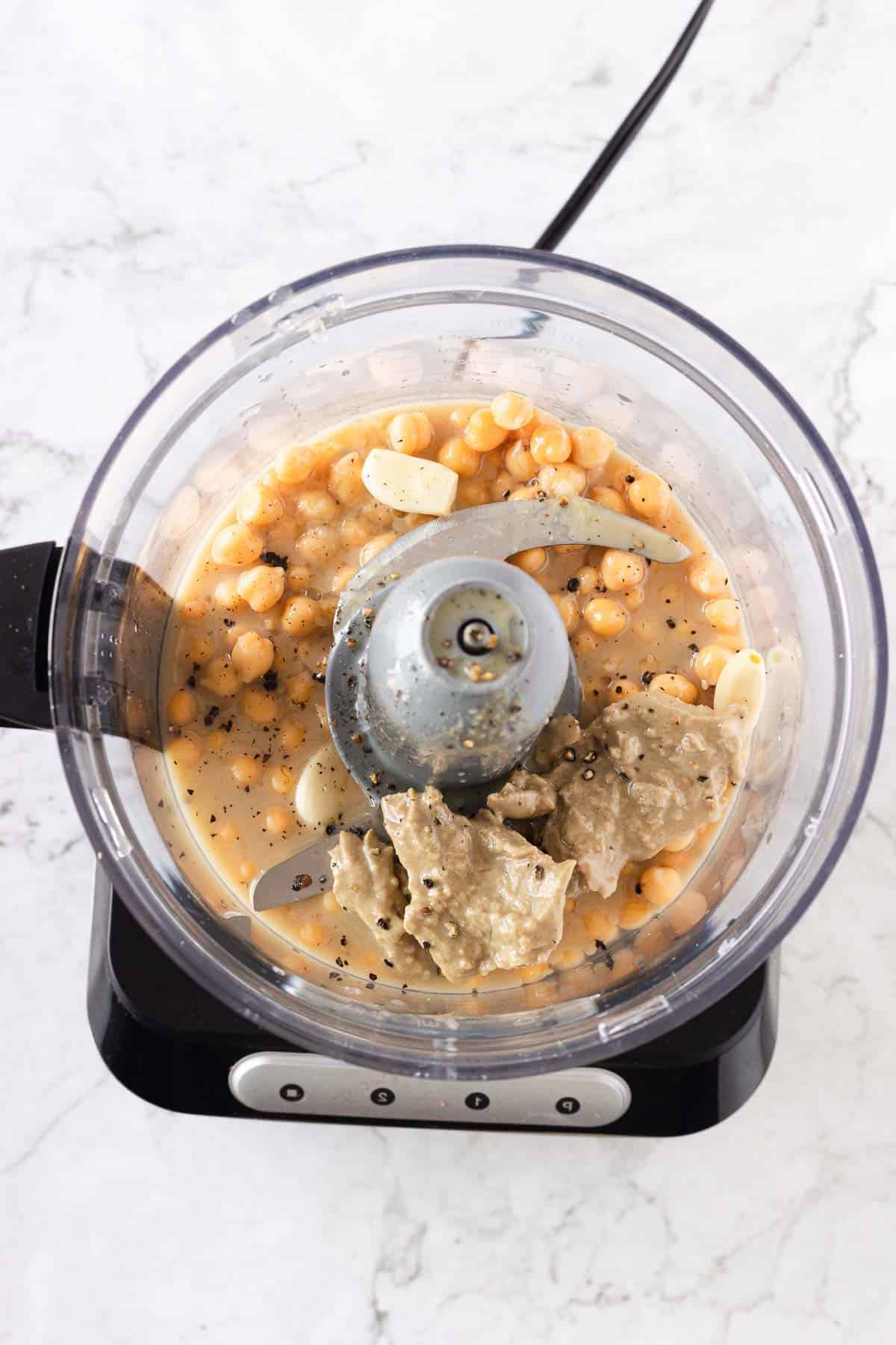 A top-down view of a food processor containing chickpeas, garlic cloves, tahini, black pepper, and liquid, presumably for making hummus. The food processor is on a marble countertop.