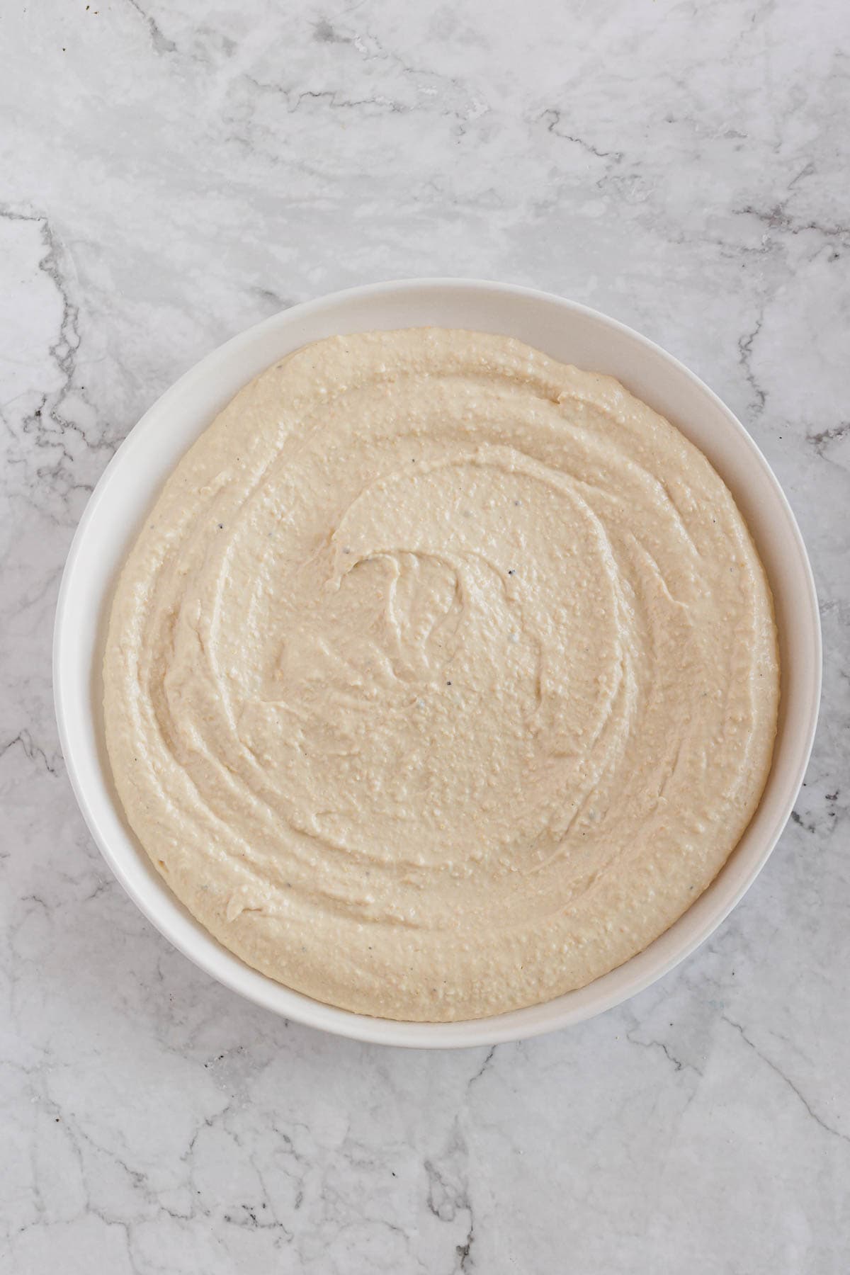 A white bowl filled with creamy beige hummus is placed on a white and gray marbled surface. The hummus has a smooth texture with slight swirls on the top.