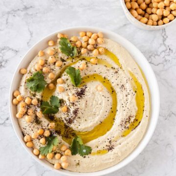 A bowl of hummus is garnished with whole chickpeas, olive oil, herbs, and spices on top. The bowl is placed on a marble countertop. In the background, there is a small bowl containing additional whole chickpeas.