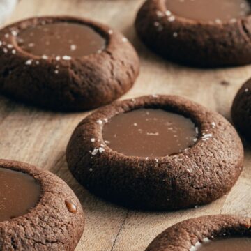 Chocolate thumbprint cookies filled with caramel are arranged on a wooden surface. These good-for-you cookies boast a glossy caramel center, topped with a sprinkle of salt. A cloth napkin is partially visible on the side, reminiscent of childhood favorites that taste better than ever.