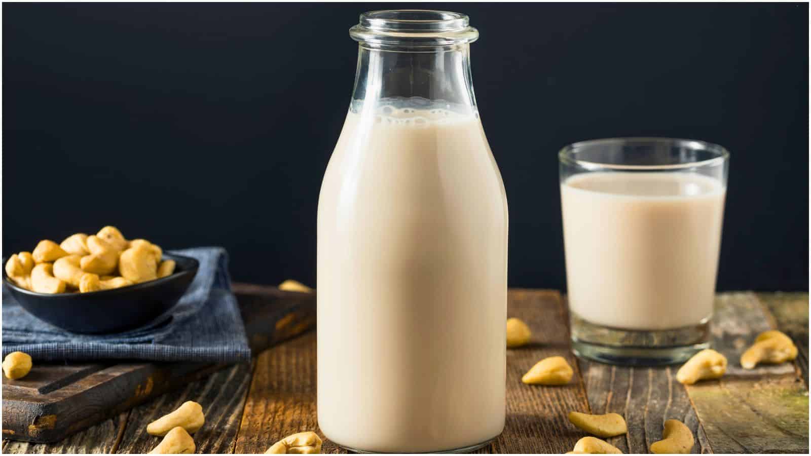 A tall container of cashew milk, with a smaller glass of cashew milk and cashews littered in the background