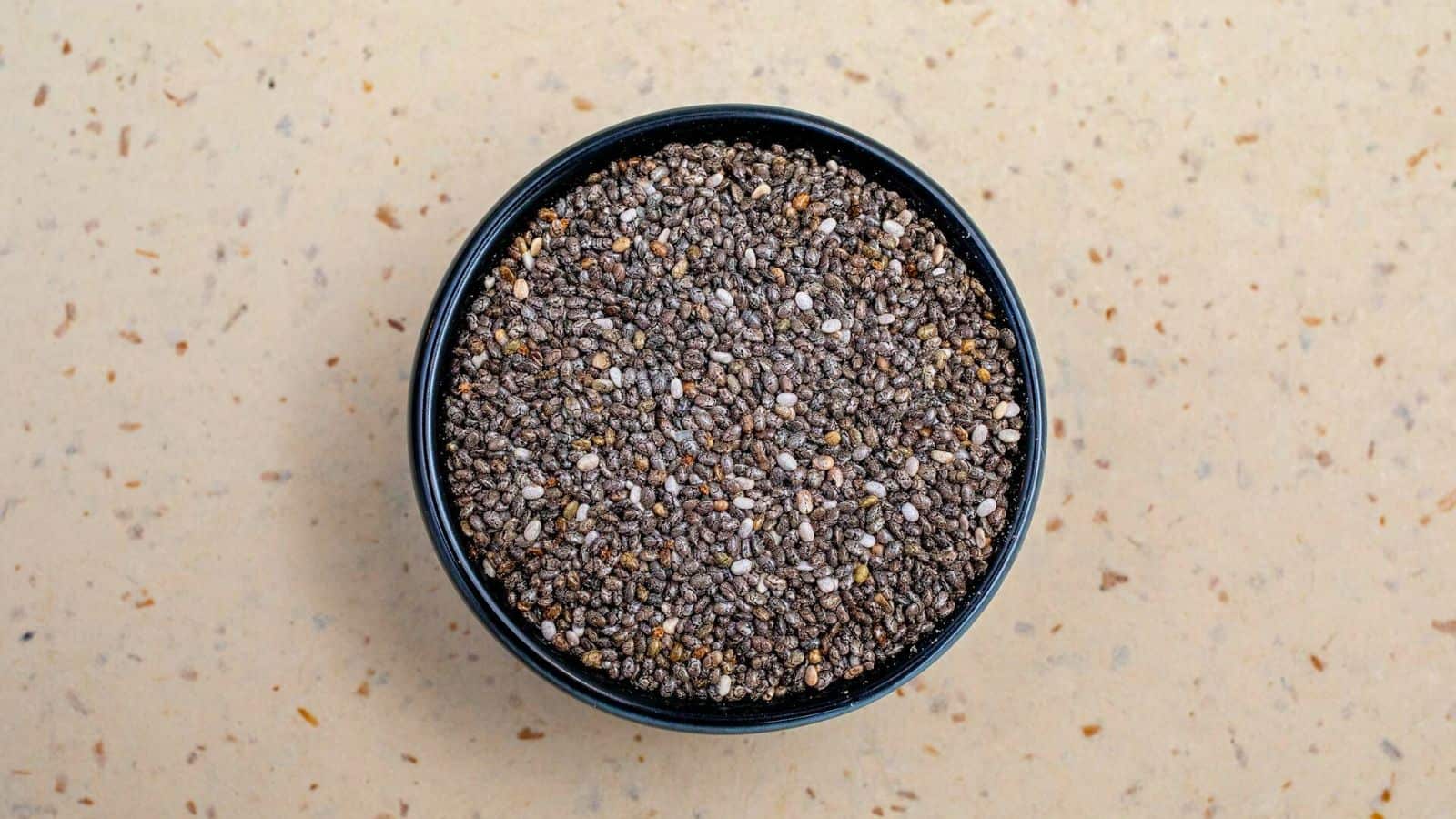 A black bowl filled with chia seeds is placed on a light beige speckled surface. The seeds are a mix of black, brown, and white colors. The image is taken from an overhead perspective.